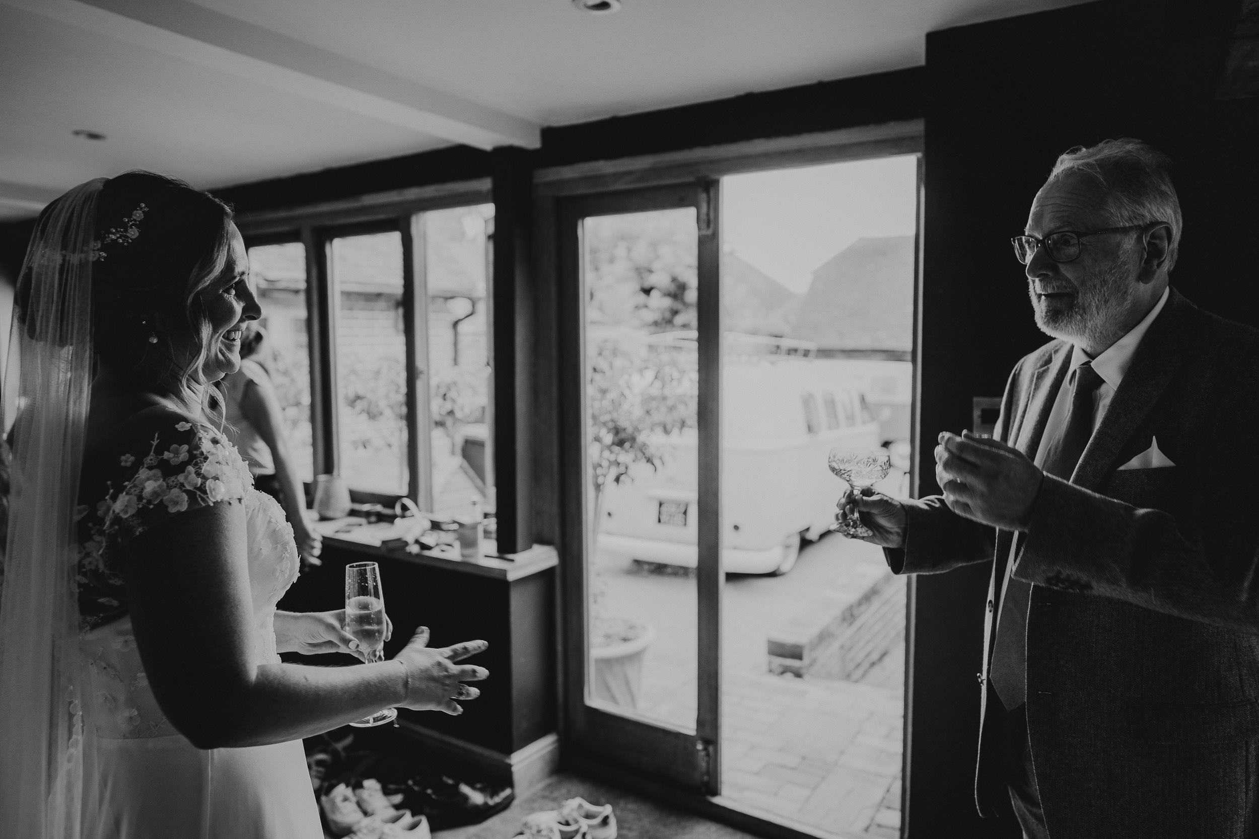 A bride and an older man engaged in conversation while holding drinks in a dimly lit room.