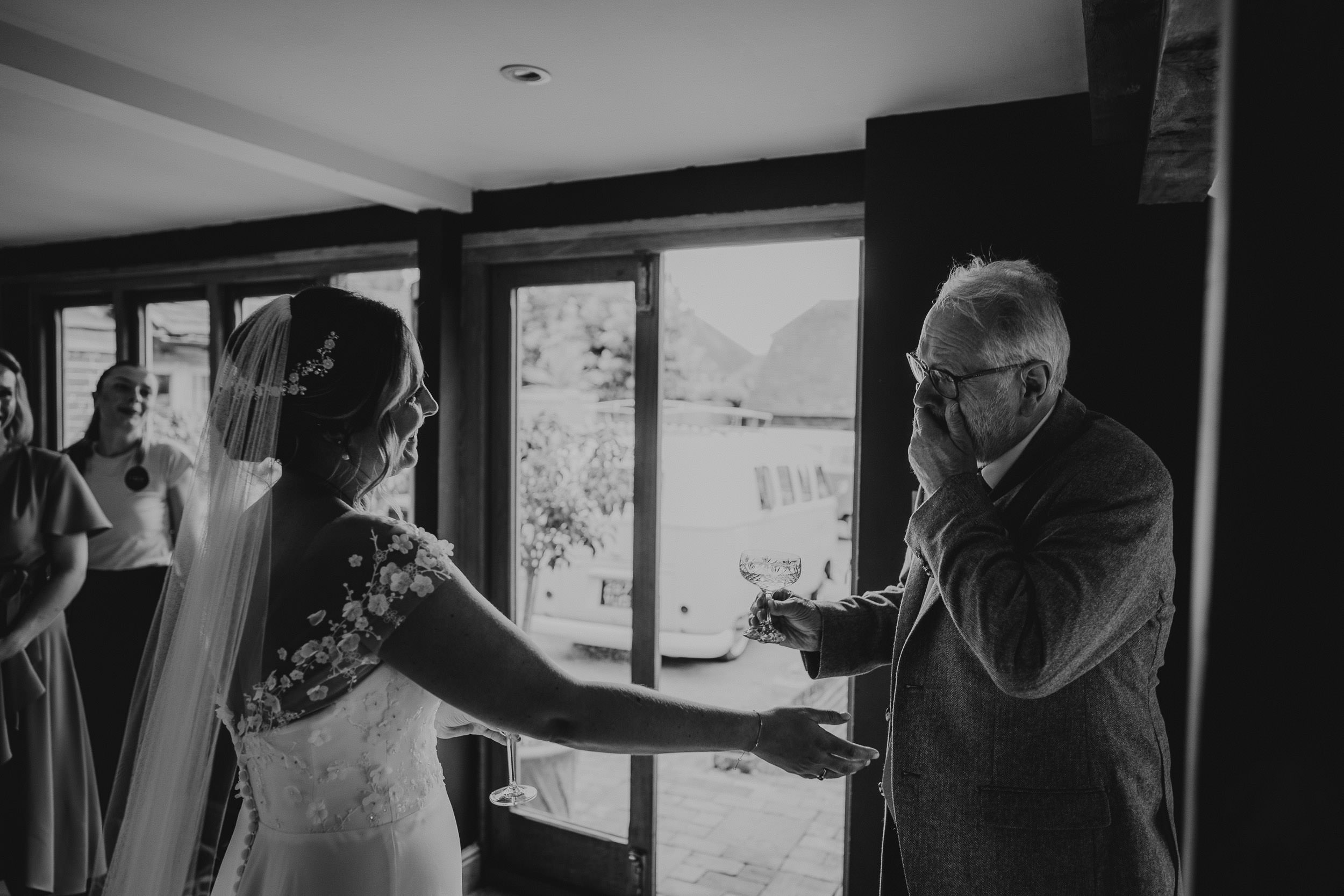 Bride in a dress and veil shares an emotional moment with an older man indoors, both holding glasses. The man appears to be wiping tears. Others are in the background.