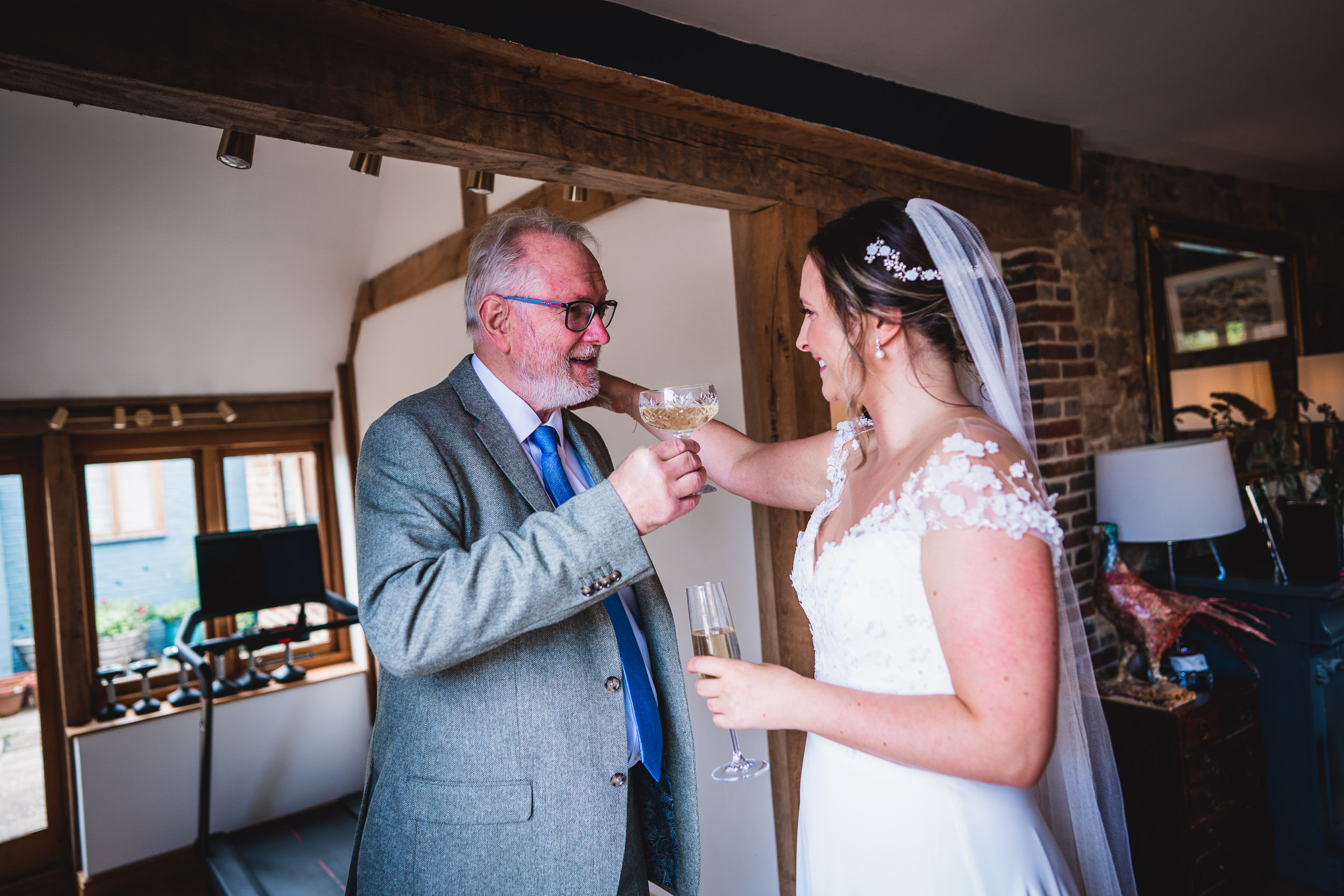 A bride in a white dress and veil shares a toast with an older man in a gray suit and blue tie inside a room with wooden beams and brick walls.