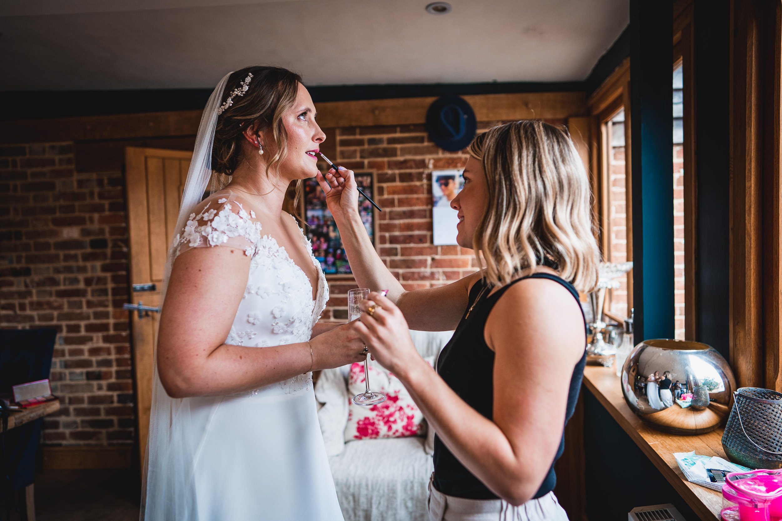 Bride having lipstick applied by a woman in a room with brick walls and large windows.