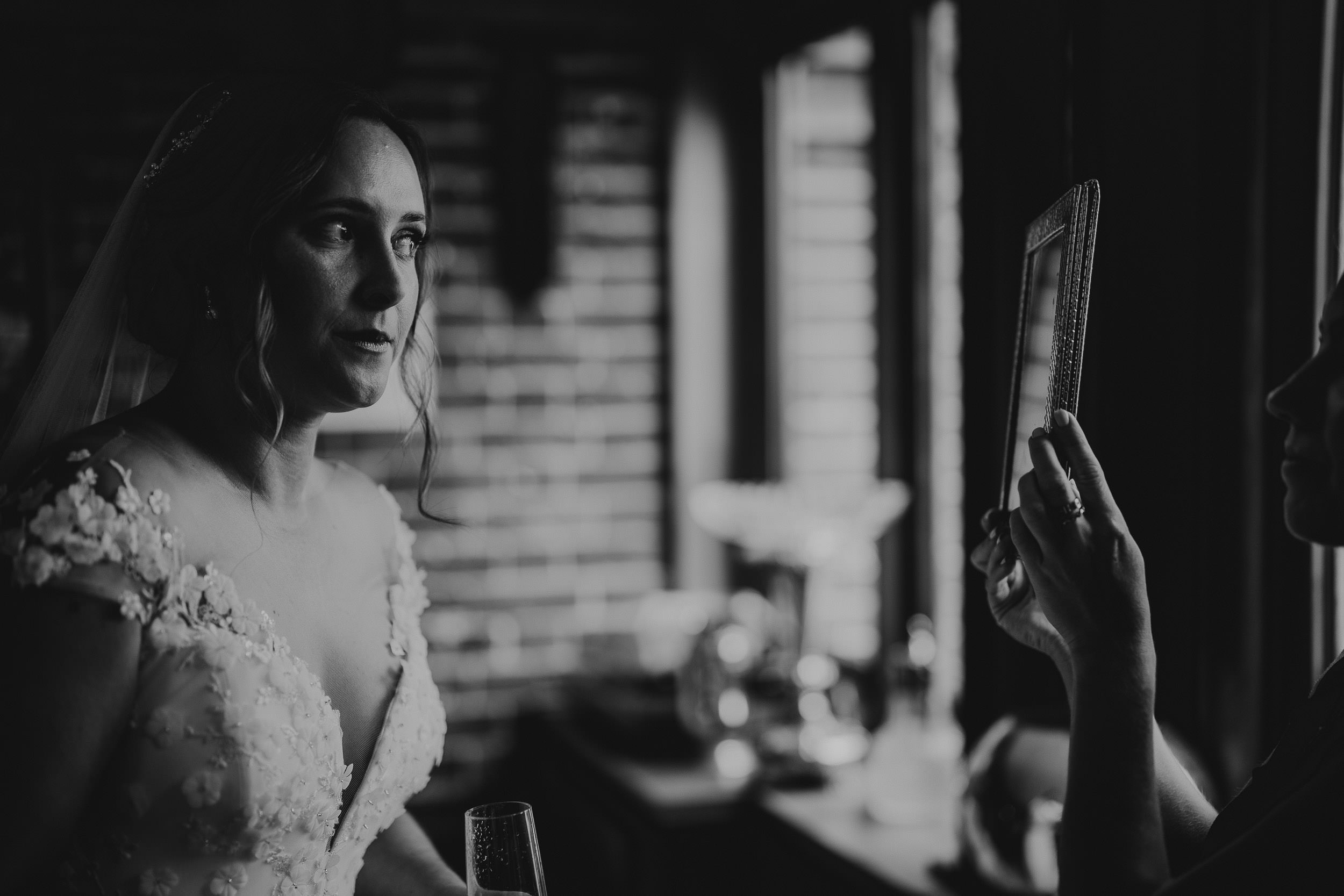 Bride in a detailed gown stands indoors, looking towards a person holding a framed object, with brick walls and soft lighting in the background.