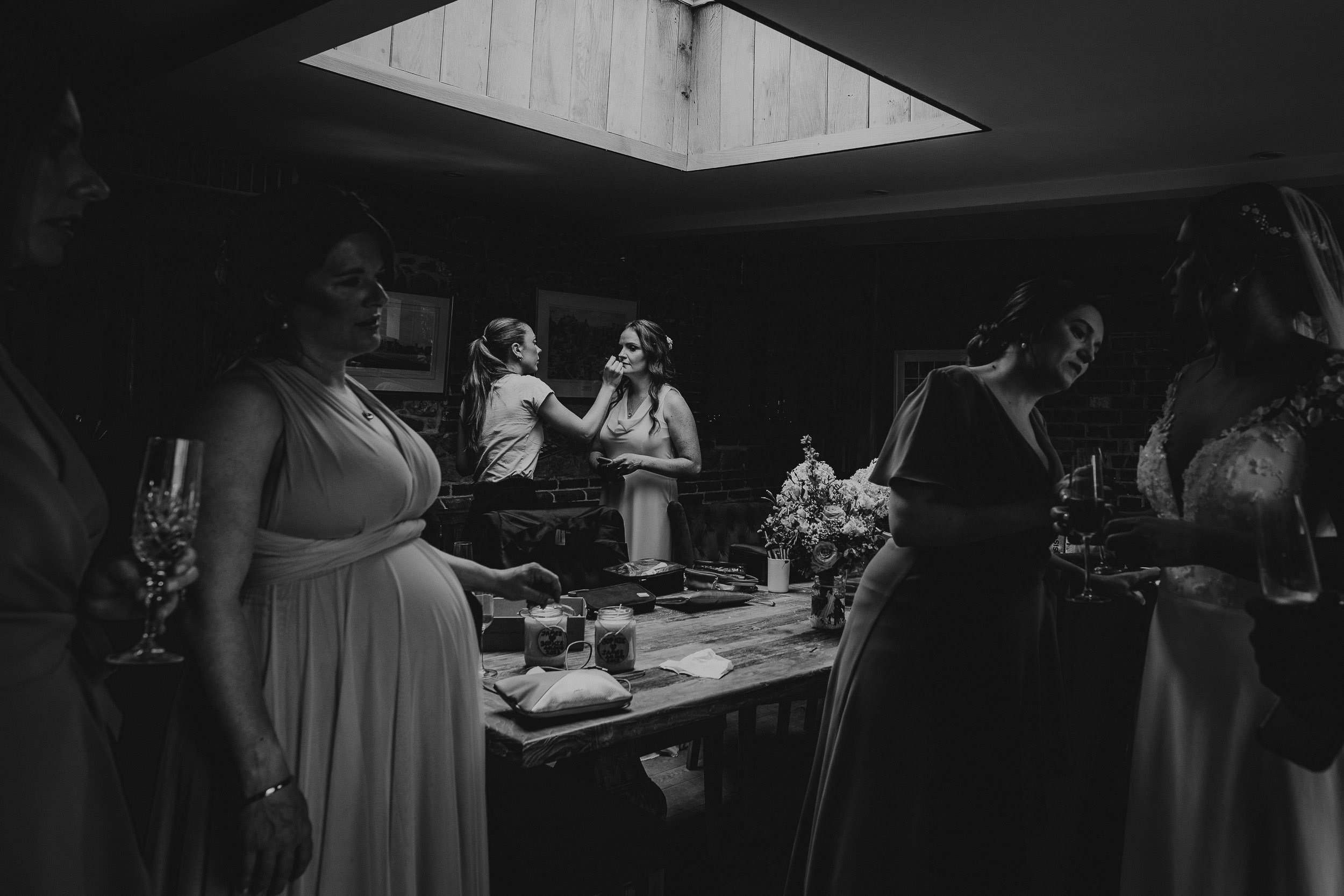 Bridesmaids in dresses interact indoors, with two embracing in the background. The room is dimly lit with a skylight above.