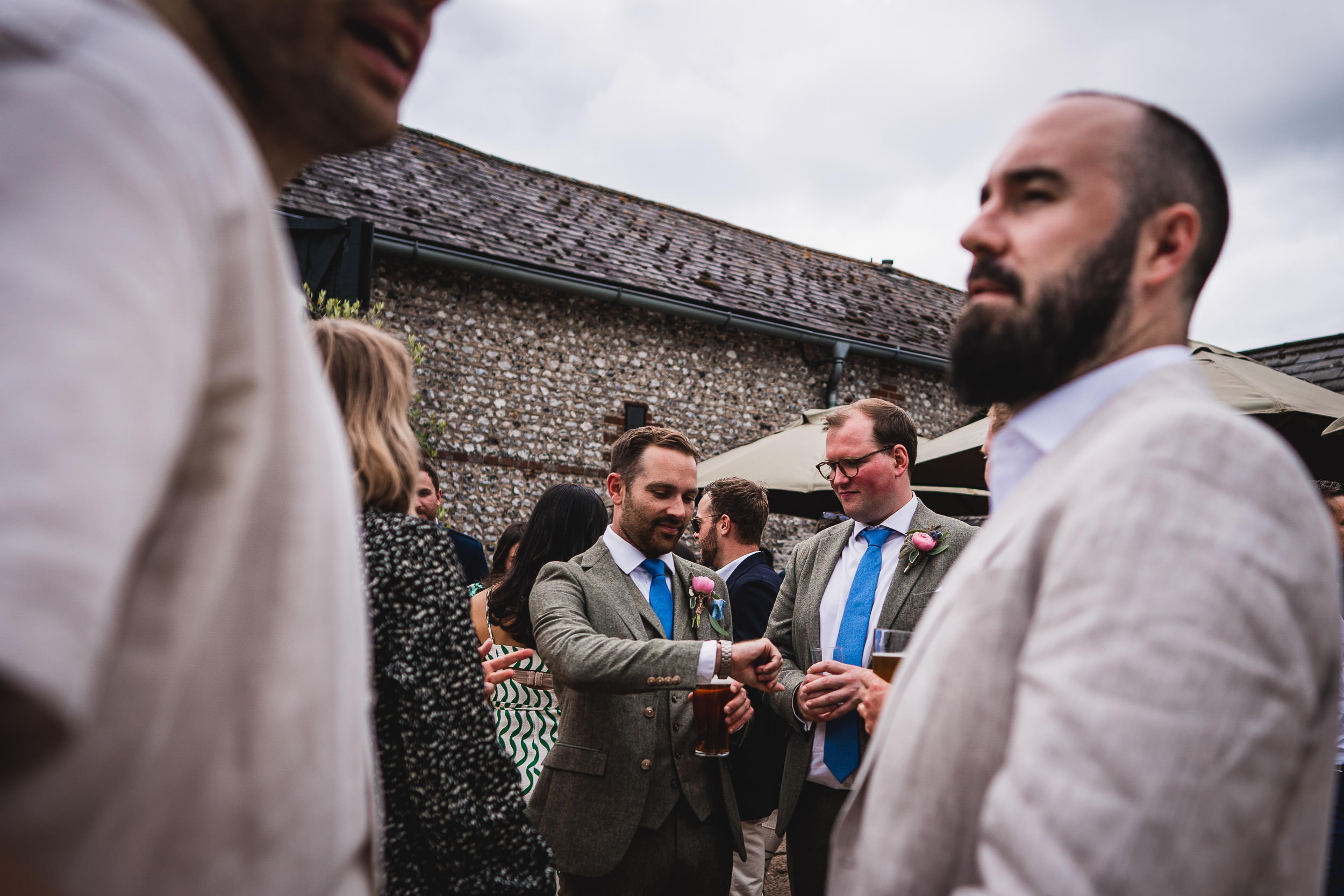 A group of people in formal attire socialize outdoors near a stone building, with some holding drinks.