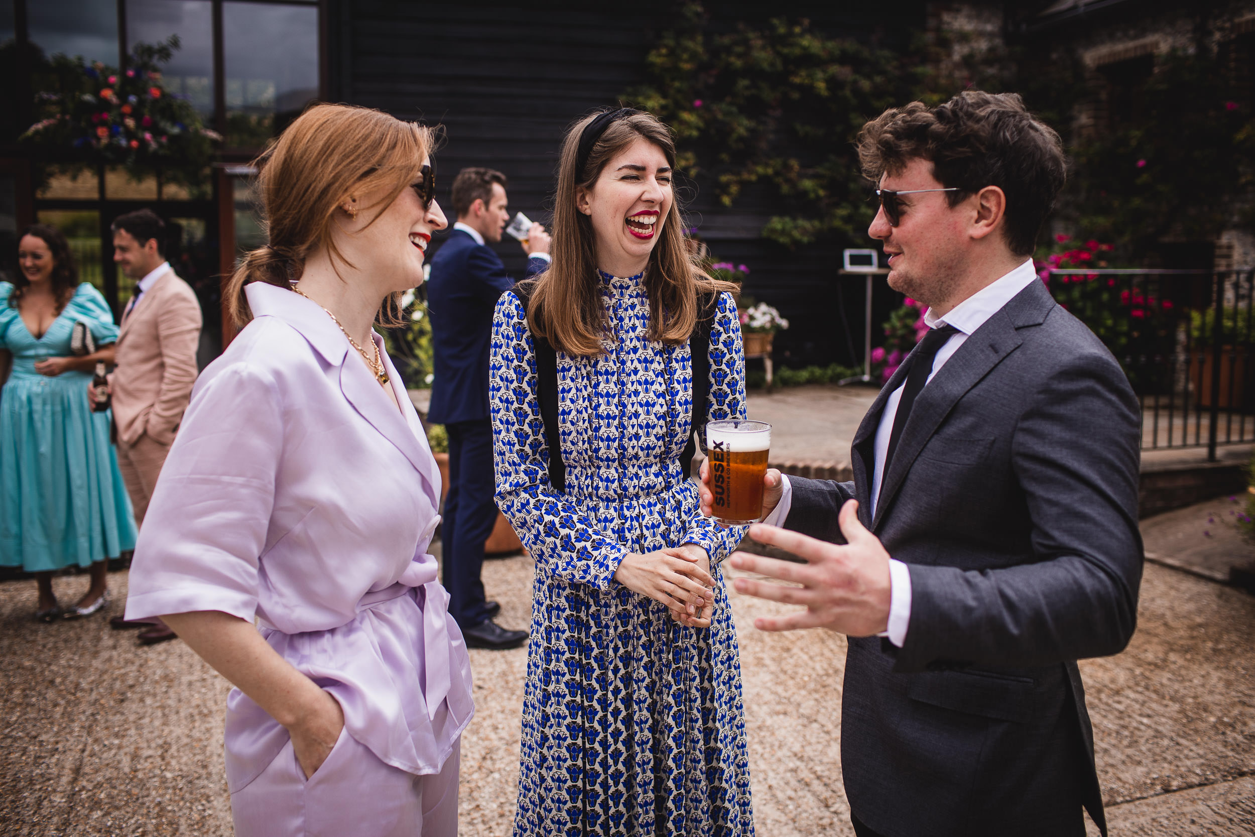 Three people are talking and laughing outside. One woman holds a drink, and the man wears sunglasses. Other people are visible in the background.