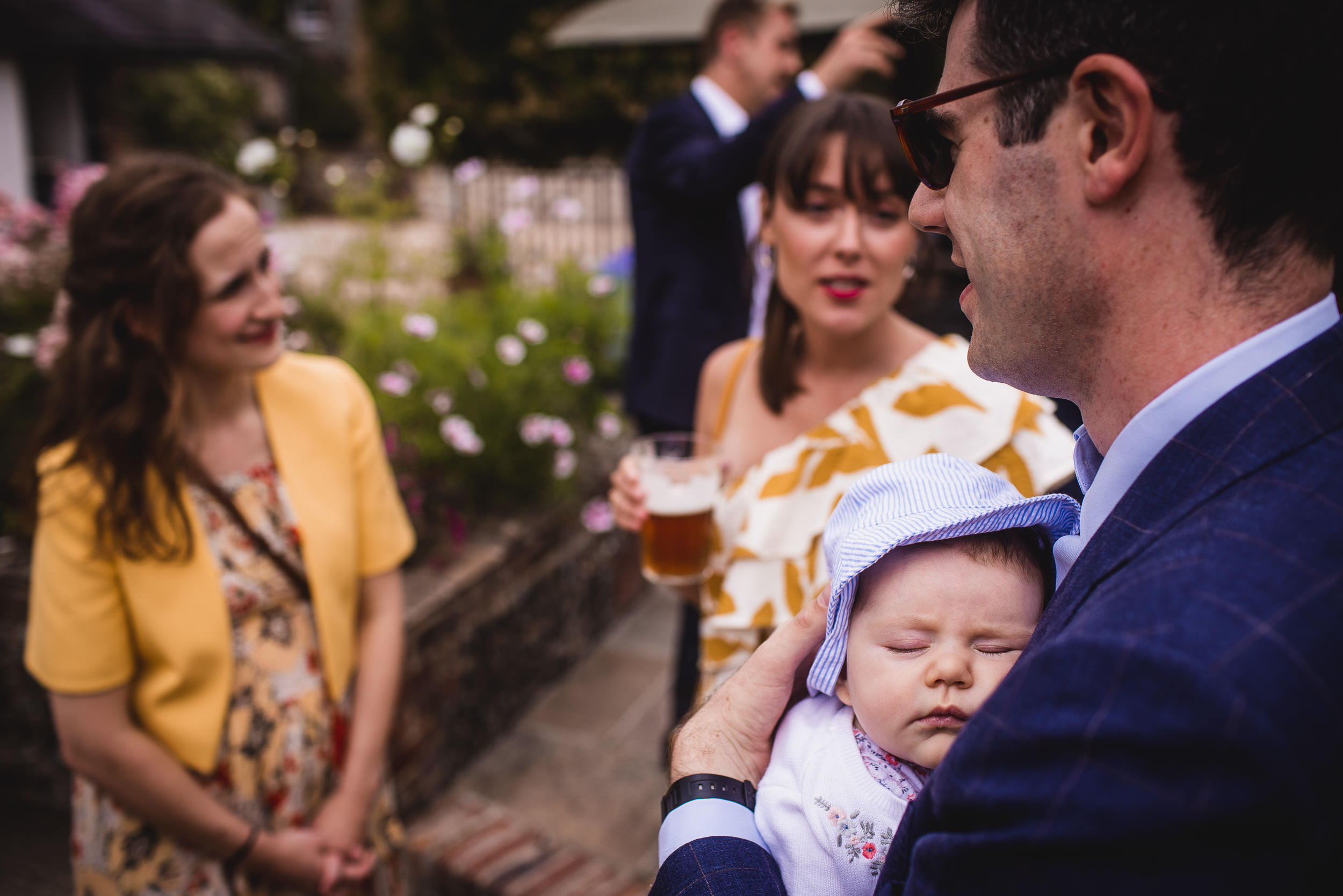 A man holds a sleeping baby while talking to two women at an outdoor gathering. One woman wears a yellow jacket, and the other holds a drink. People are in the background.