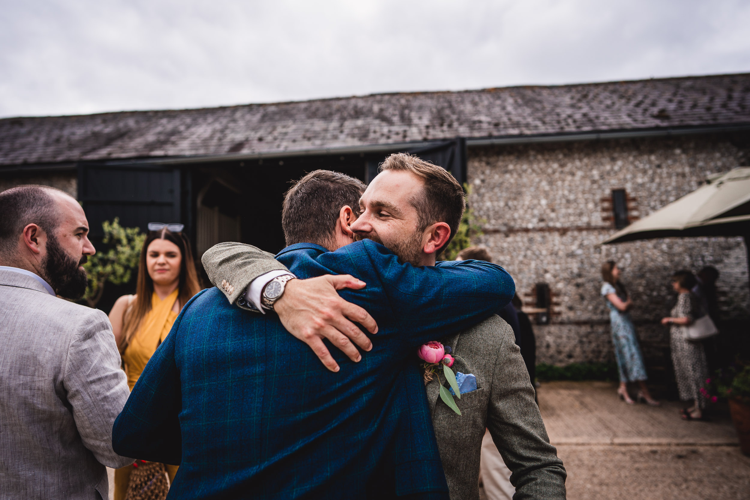 Two men hugging at an outdoor event, surrounded by a few people. A barn-like building is in the background with cloudy skies above.