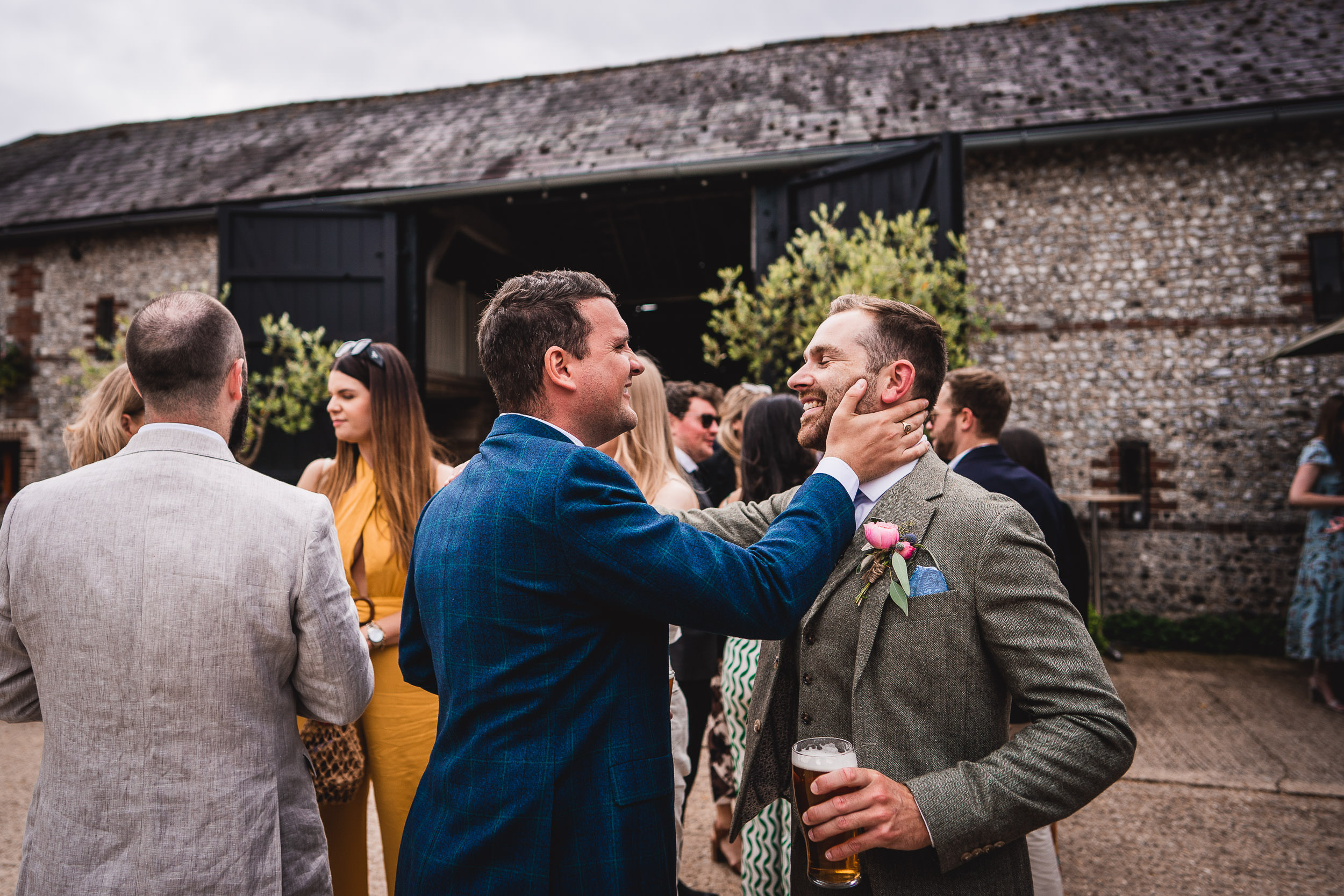 Two men in suits share a joyful moment at an outdoor gathering. One touches the other's face, while holding a drink. Several people are in the background near a rustic building.