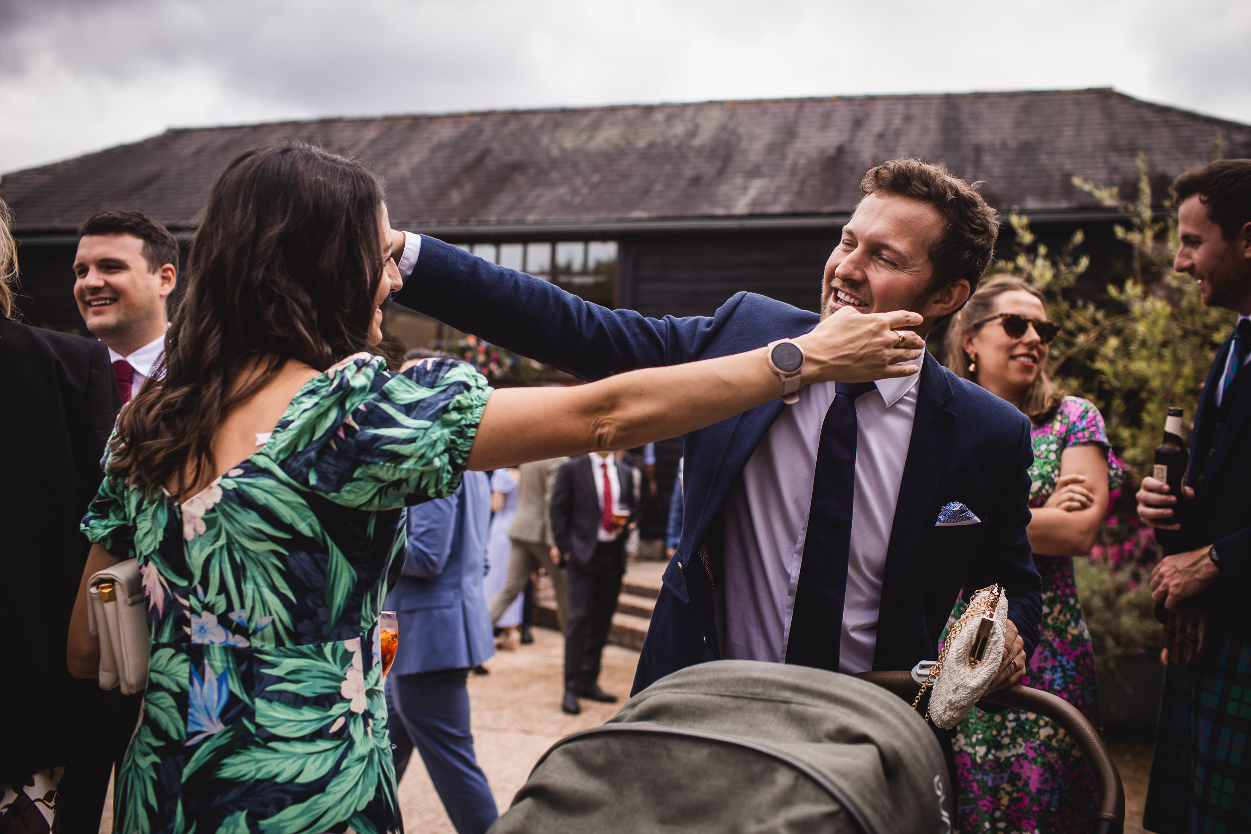 A man in a suit playfully interacts with a woman in a floral dress at an outdoor gathering. A baby stroller is in the foreground, and other people are socializing in the background.