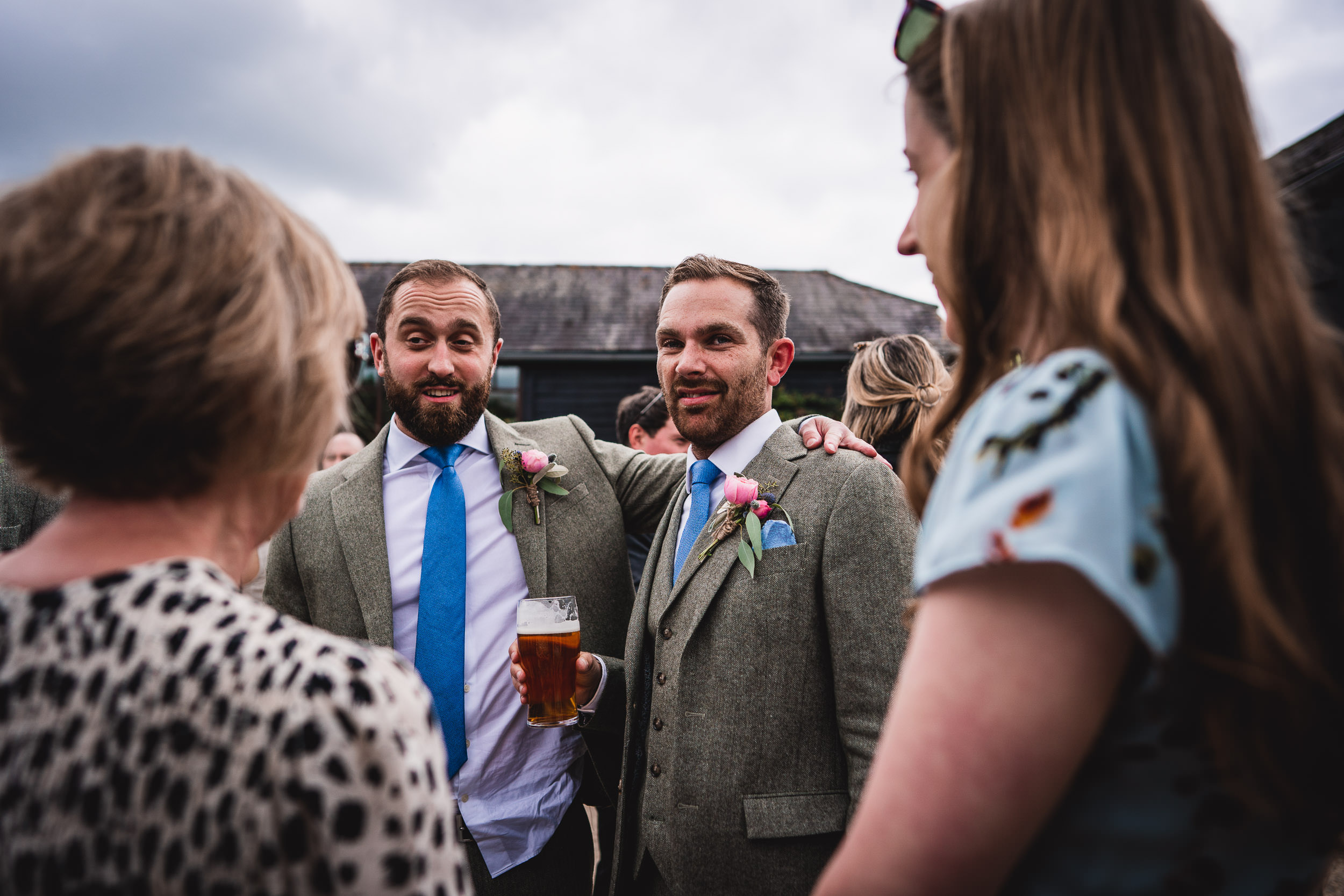 Two men in suits with floral boutonnieres share a moment outdoors; one holds a pint of beer. Other people stand around them, engaging in conversation.