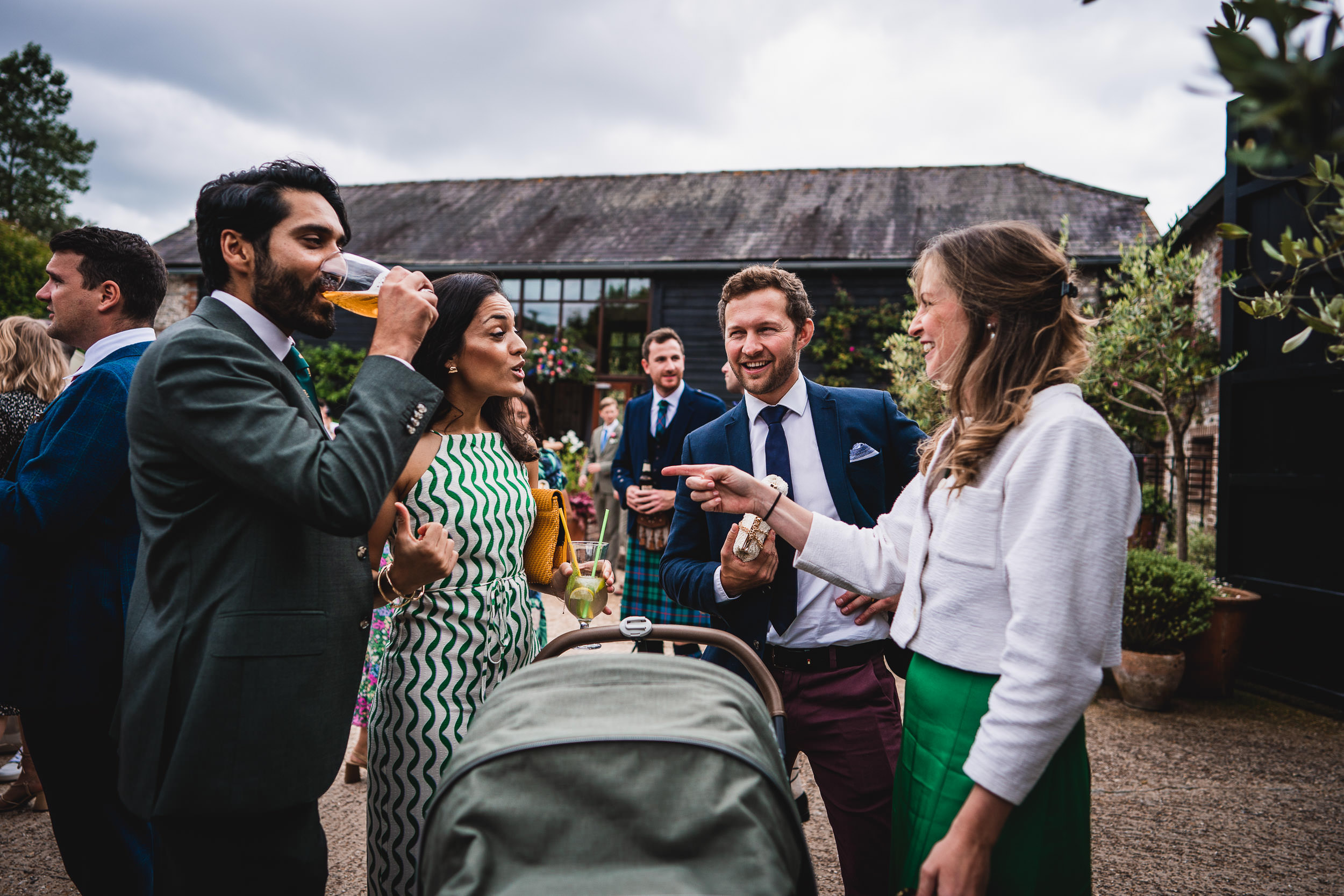 A group of people in formal attire stand outside a rustic building, talking and smiling. A woman points at a baby stroller, while others hold drinks. Overcast sky in the background.