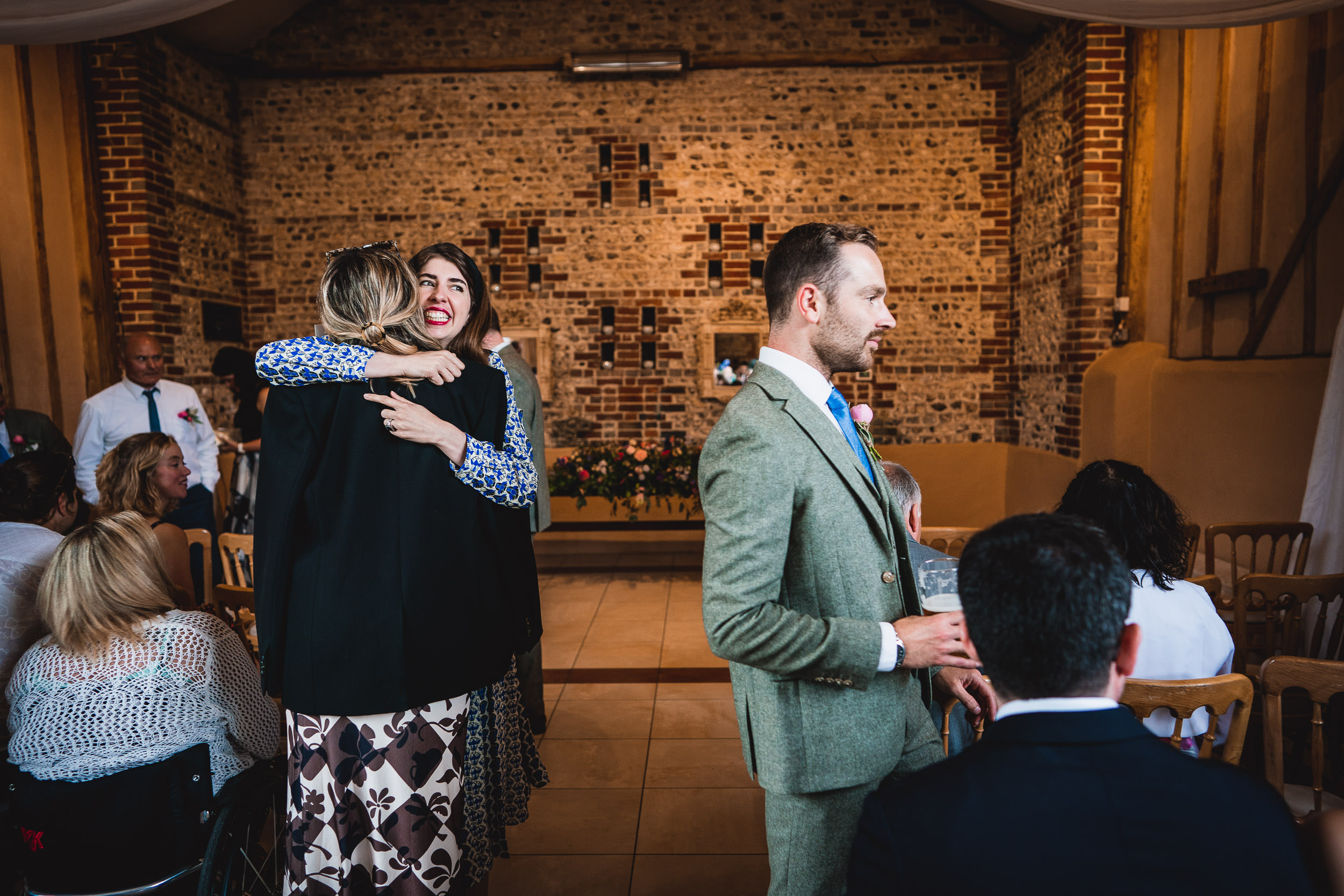 Guests in a warmly lit room interacting; a woman hugs another, while a man in a green suit stands nearby holding a glass.