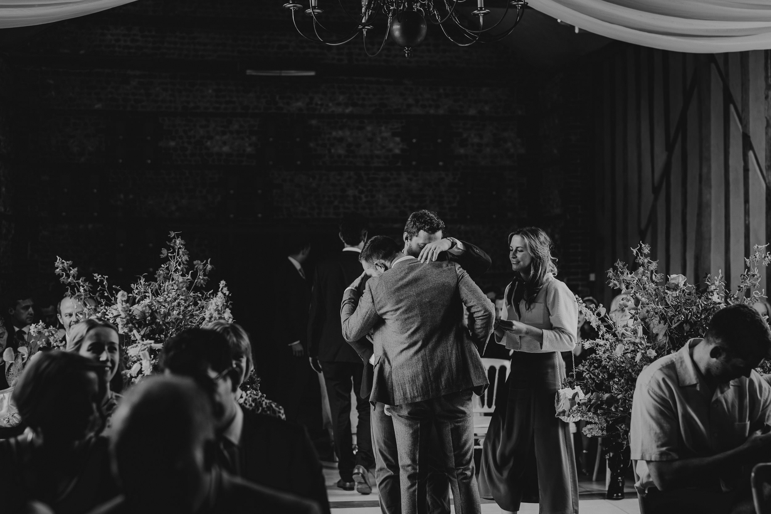 Guests embrace and chat at an indoor event, surrounded by floral arrangements and a hanging chandelier in a dimly lit room.