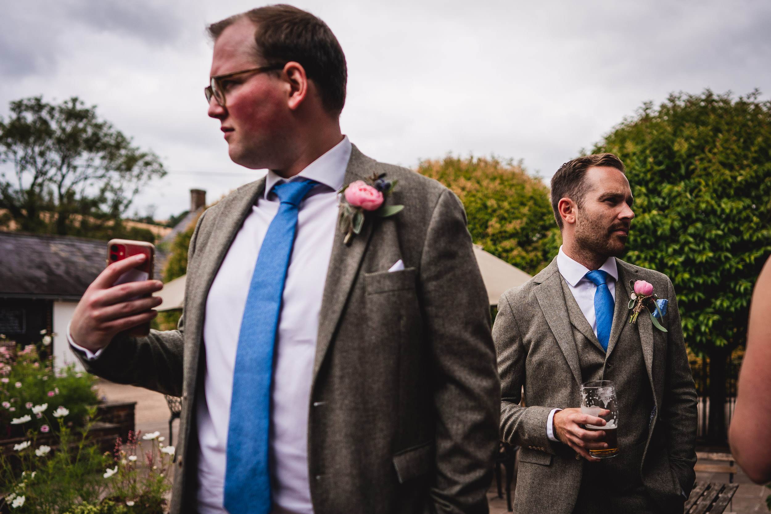 Two men in suits with blue ties, holding drinks and standing outdoors. One is holding a phone. Trees and buildings in the background.