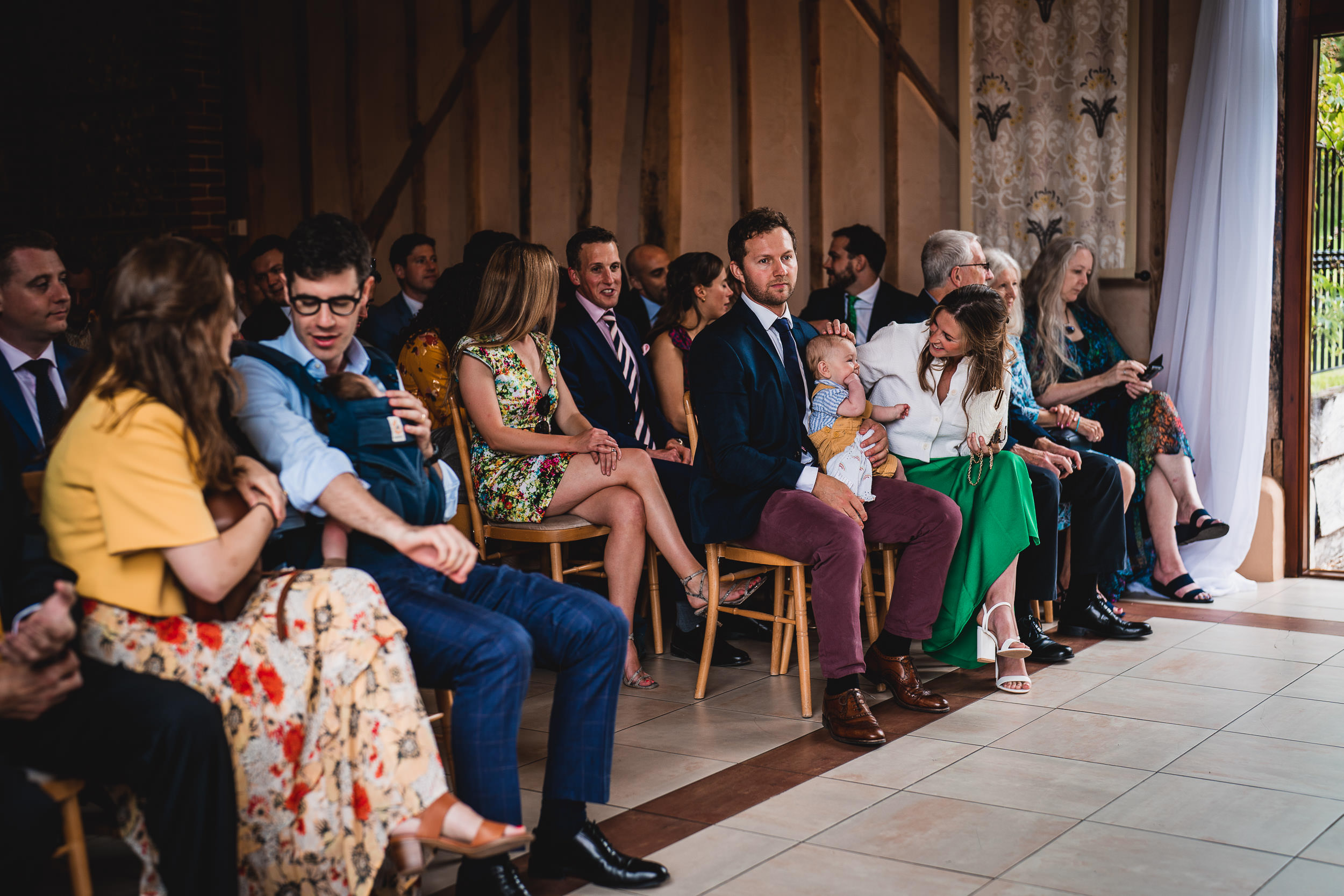 A group of people sitting on wooden chairs, including adults and children, in a decorated indoor setting. Some people are talking, while others look ahead.