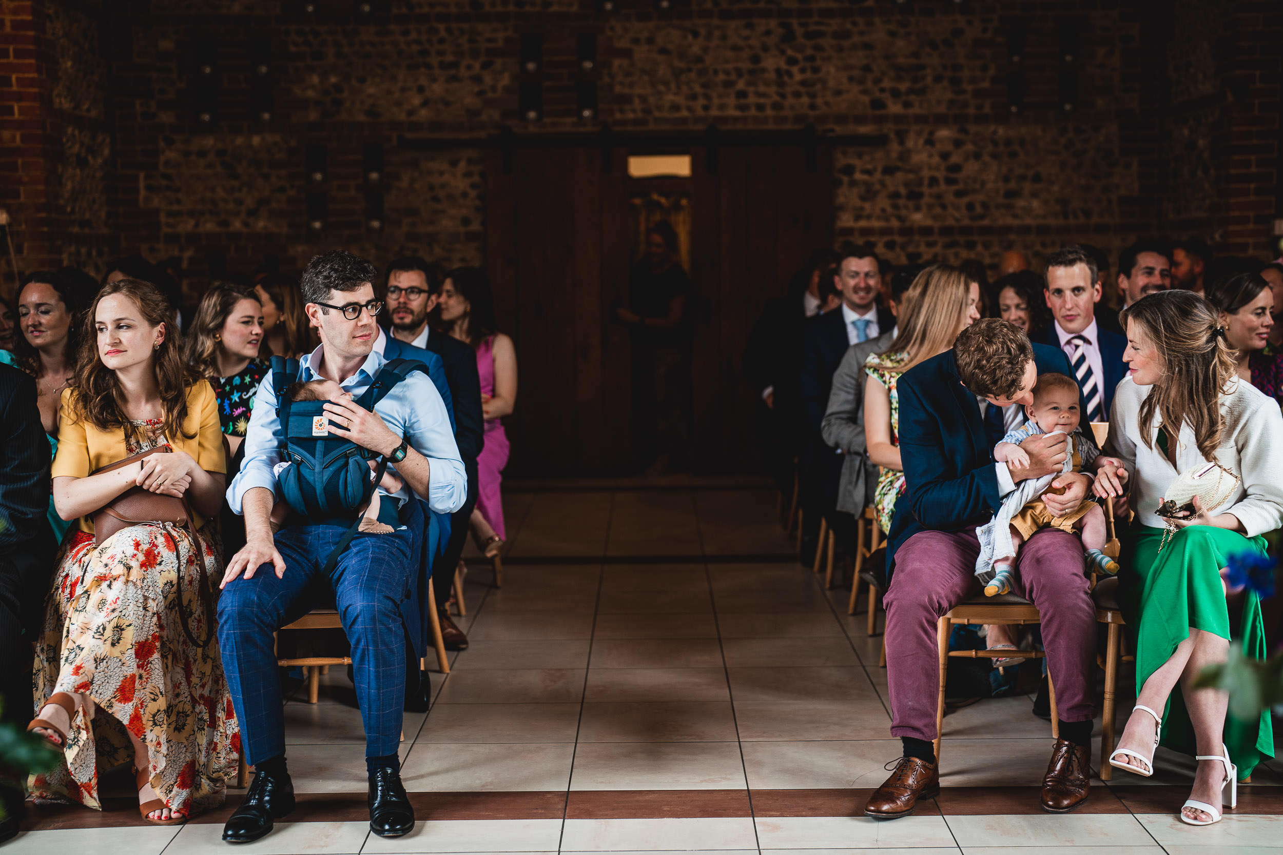 People seated in rows at an indoor event, some holding babies.