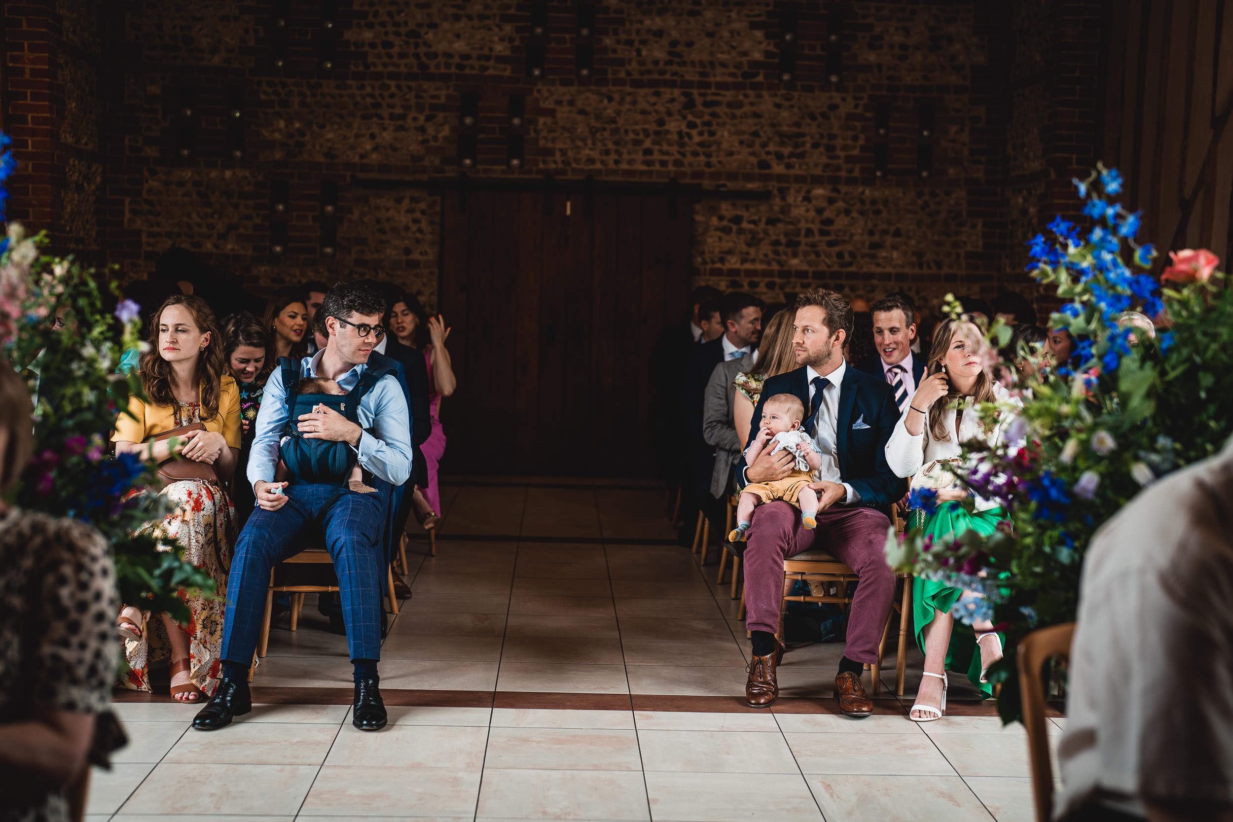 A wedding audience sits on wooden chairs, some attentive and others distracted. Bright floral arrangements decorate the aisle, and a man holds a baby.