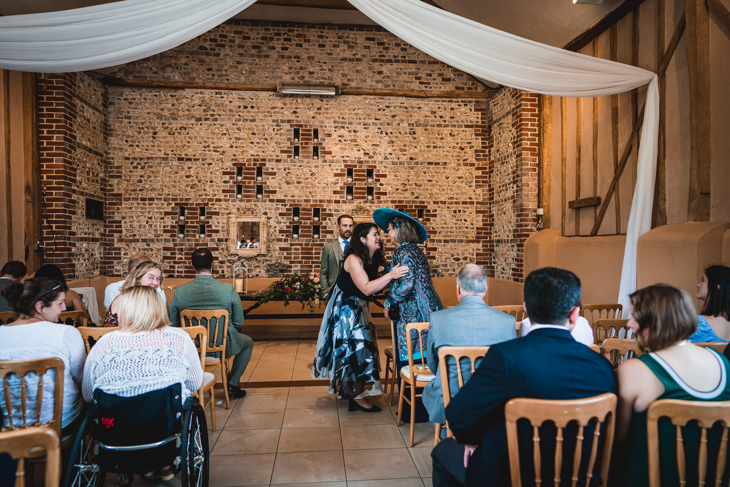 Guests sit and chat in a brick-walled venue before a ceremony. Some are dressed formally, and one person wears a large hat. Rows of wooden chairs are arranged, and a floral arrangement is visible.