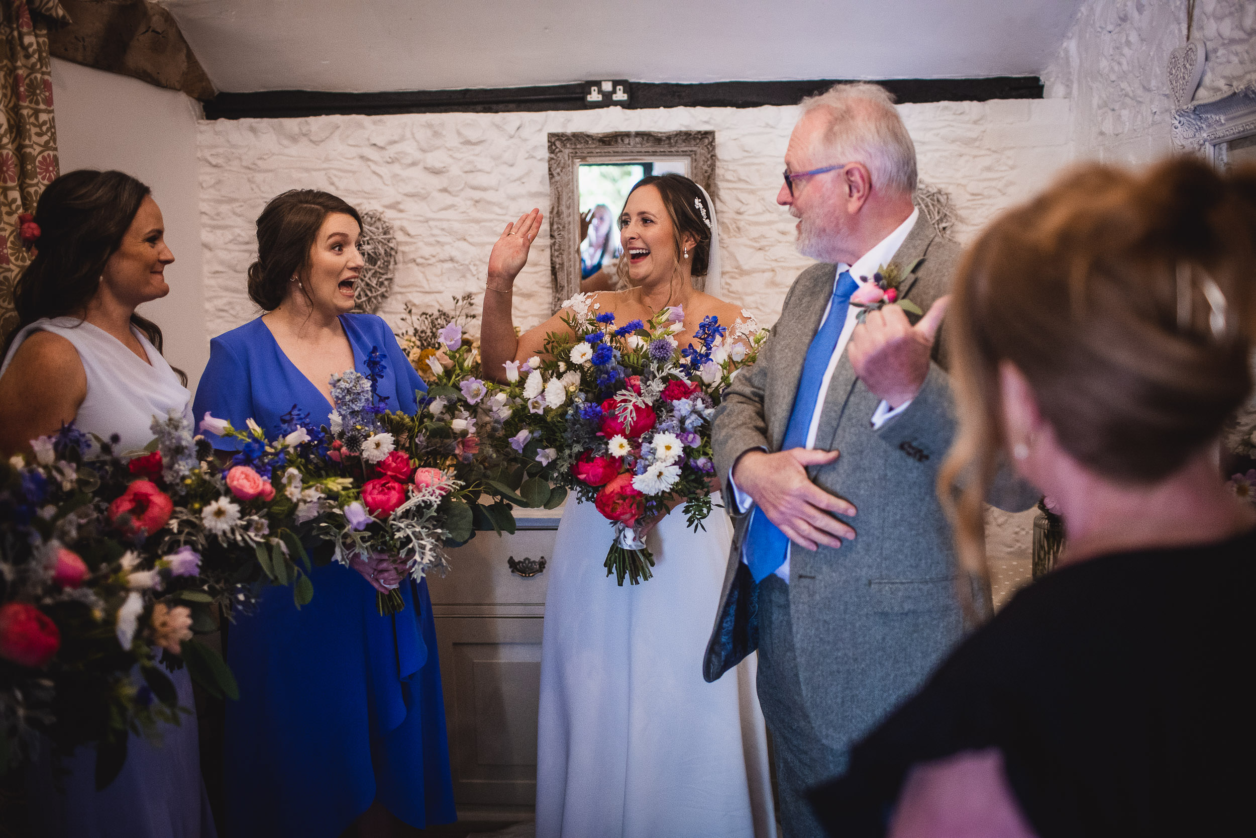 A bride and her bridal party celebrate in a room with stone walls. The women hold colorful bouquets, and a man in a suit smiles while adjusting his tie.