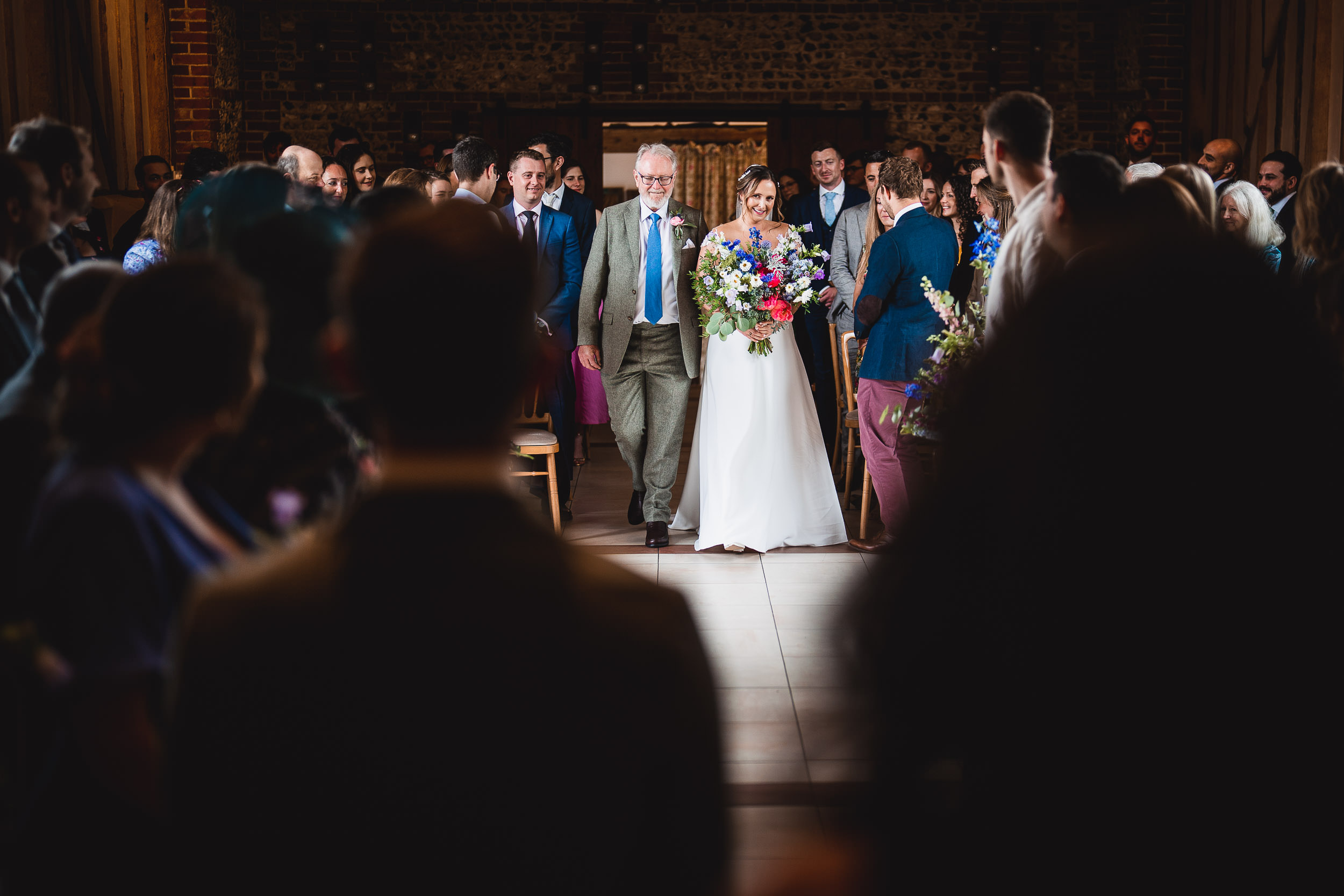 A bride in a white dress walks down the aisle with an older man in a suit, surrounded by seated and standing guests in a dimly lit venue.