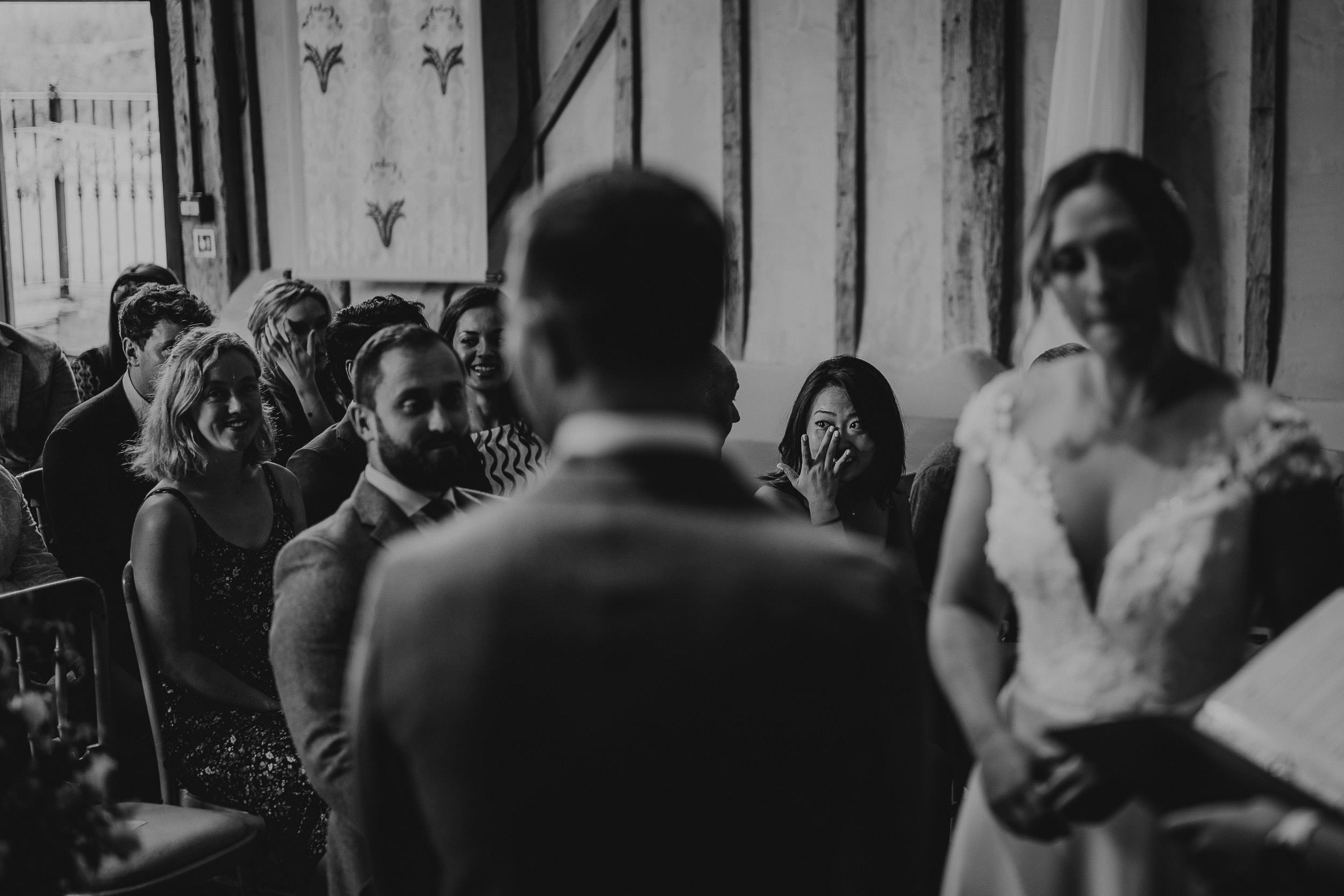 Bride and groom stand facing each other during a wedding ceremony, with guests seated and watching attentively. The setting is indoors with dim lighting.