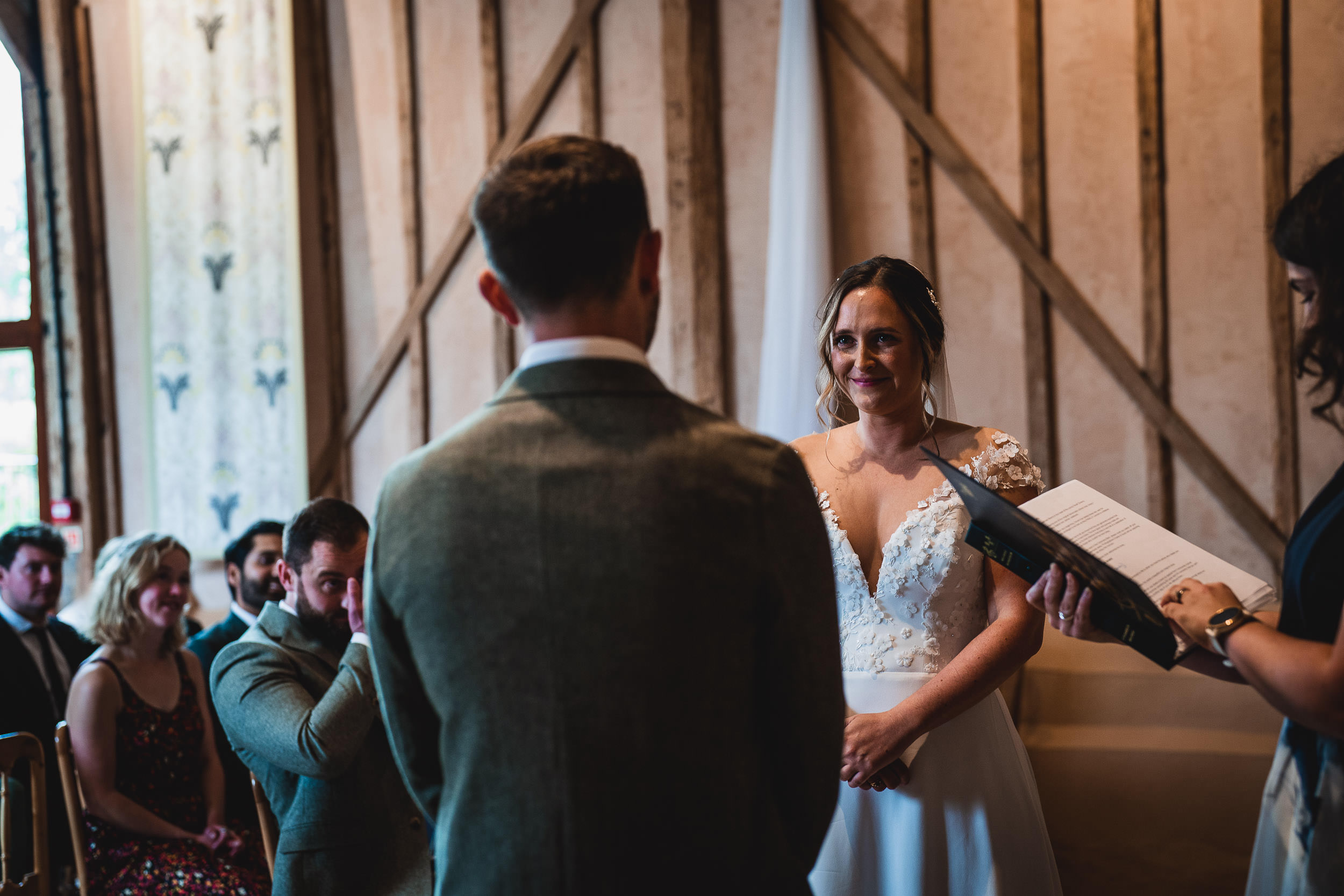 A couple stands facing each other during a wedding ceremony indoors. A person holds a book, officiating. Guests are seated nearby.