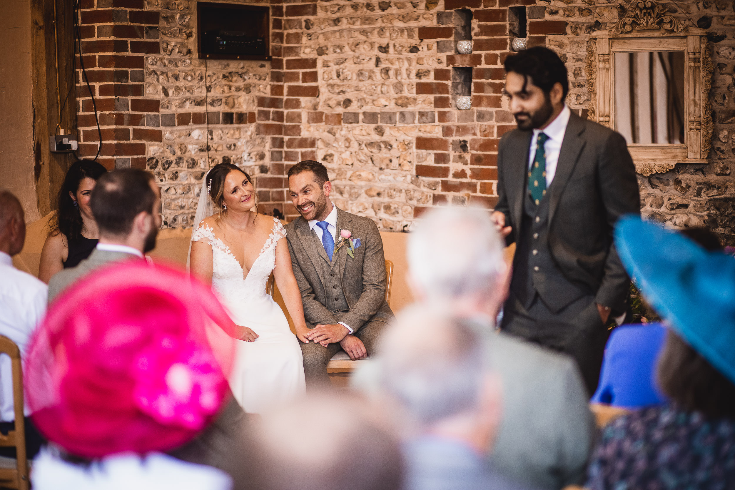 A bride and groom sit together smiling as a man in a suit speaks to an audience at a wedding ceremony in a rustic brick interior.