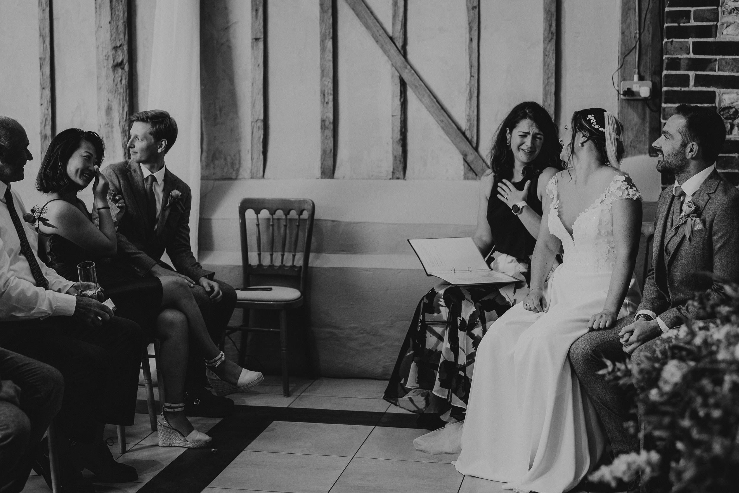 A bride and groom sit together during a ceremony, with guests seated nearby. The setting has rustic wooden beams and a brick wall. Black and white photo.