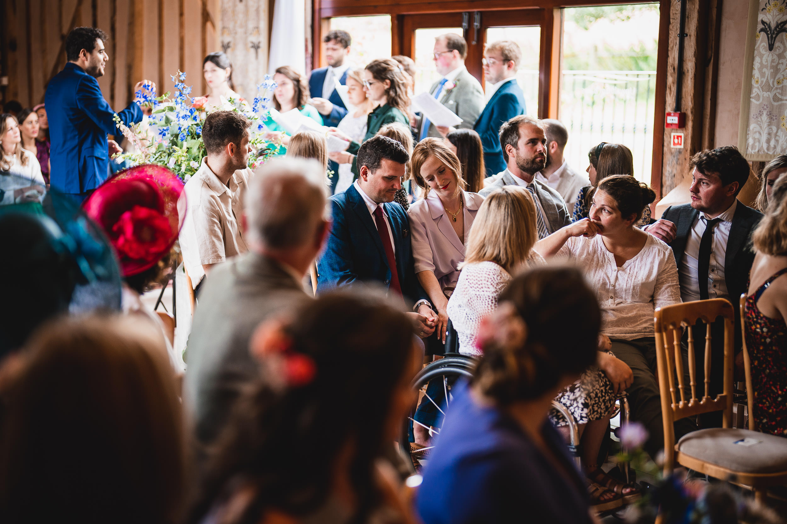 A group of people seated at a gathering, some wearing formal attire. They are indoors with some guests standing, engaging and conversing.