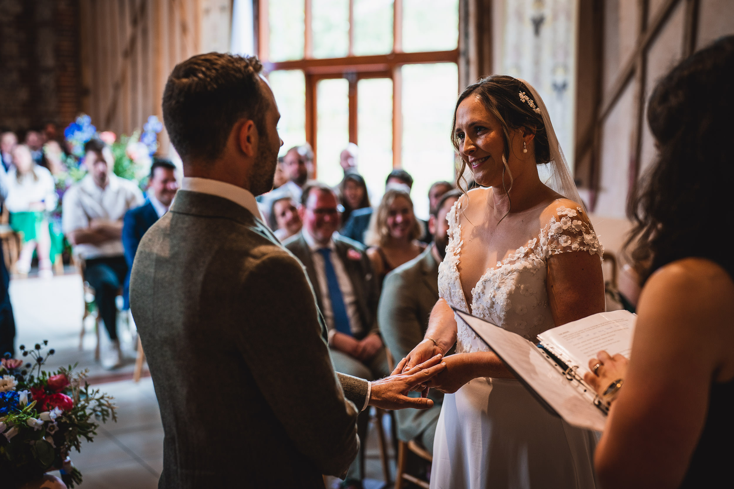 A couple exchanges vows during a wedding ceremony, with guests seated in the background and natural light streaming in through a large window.