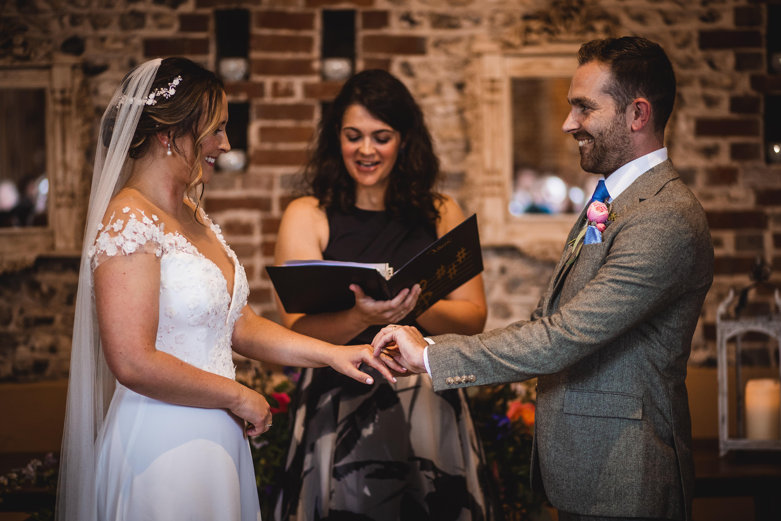 A couple exchanges rings during a wedding ceremony, with a person officiating in the background. The bride wears a white dress and veil, and the groom wears a suit.