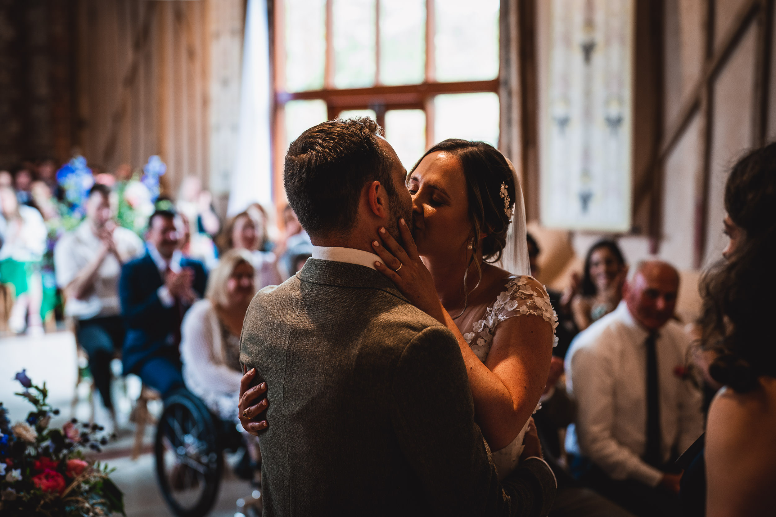 A bride and groom share a kiss in a warmly decorated room, surrounded by applauding guests.