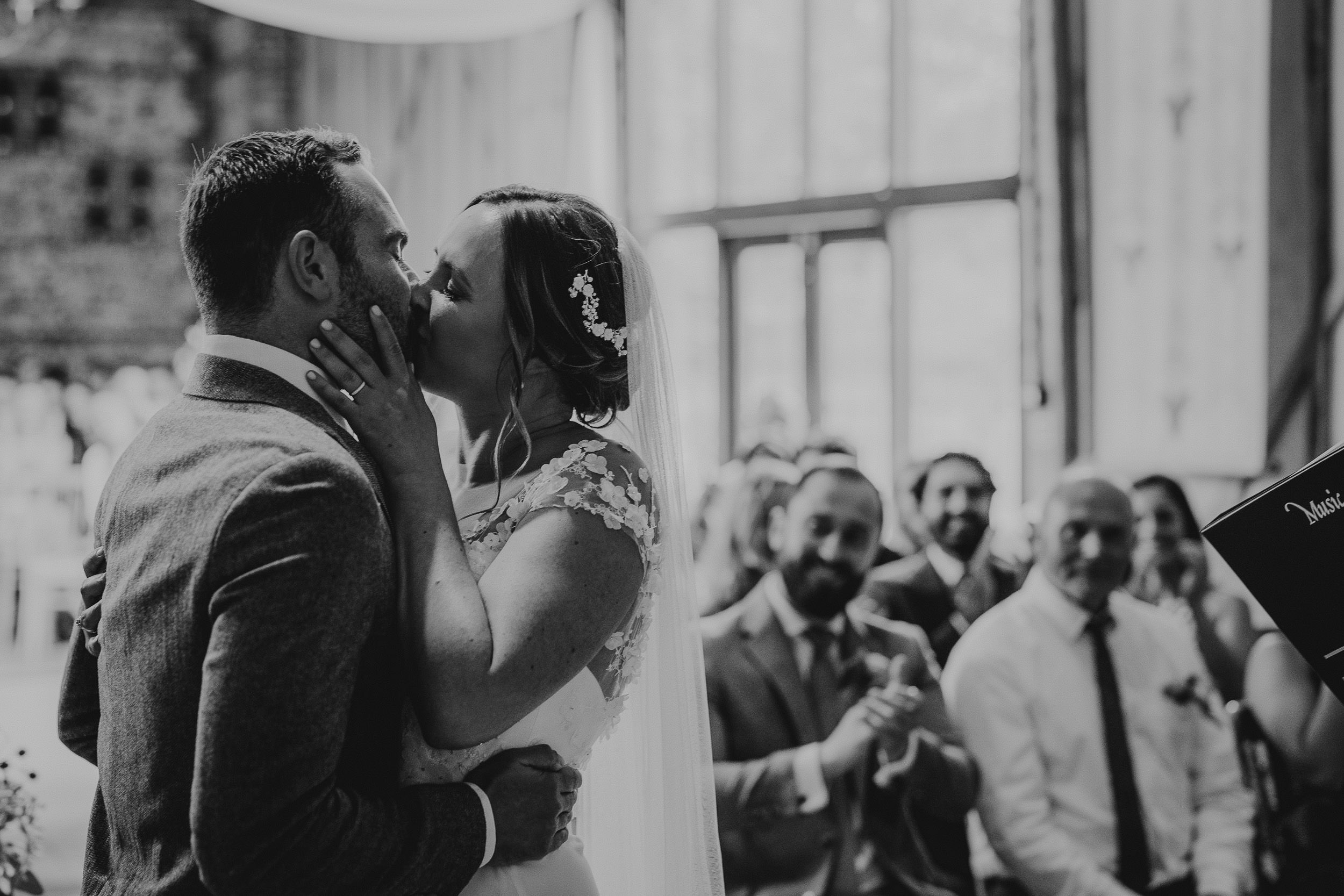 Bride and groom share a kiss during their wedding ceremony. Guests in the background watch and clap.