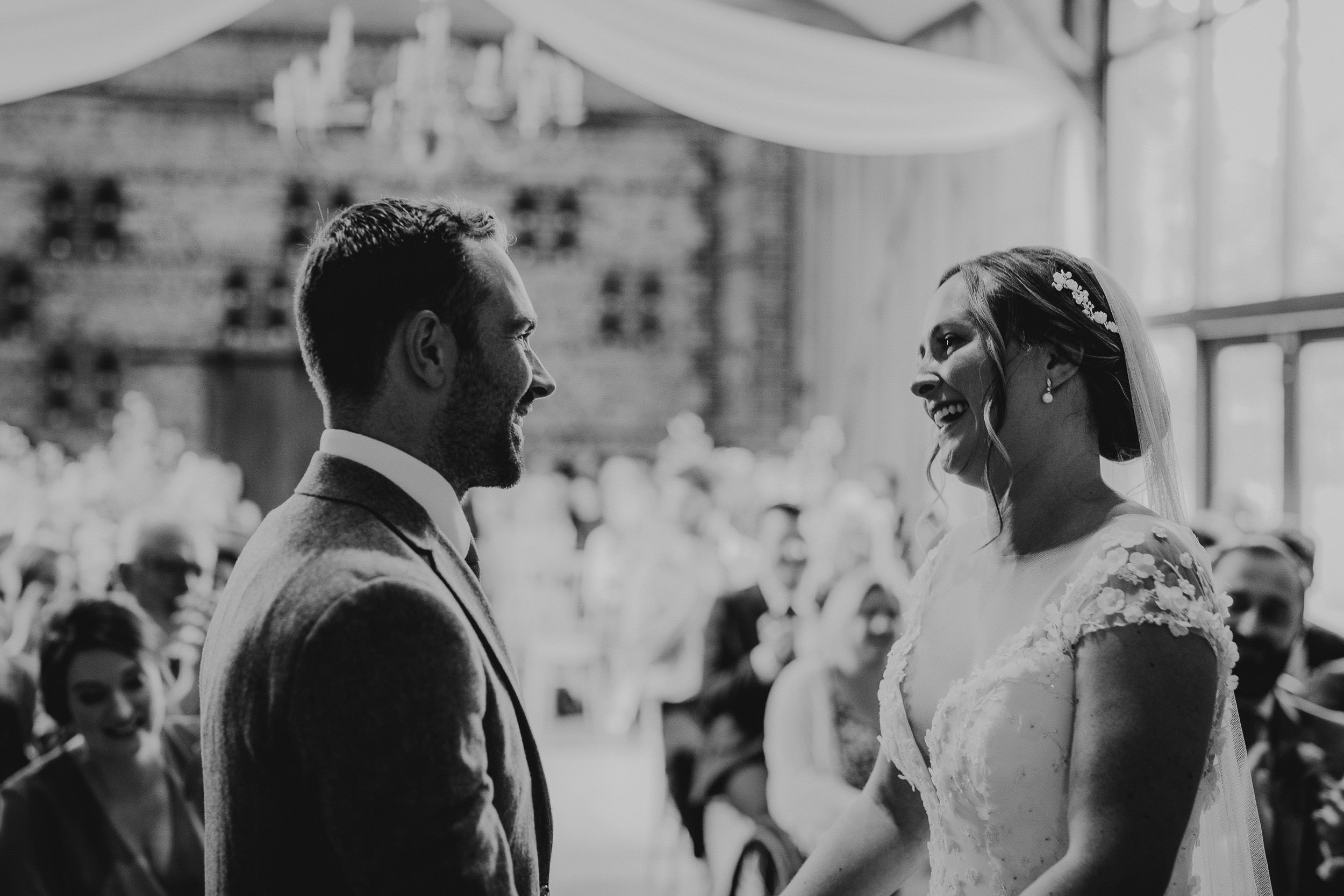 Bride and groom smile at each other during a wedding ceremony in a hall with guests seated behind.