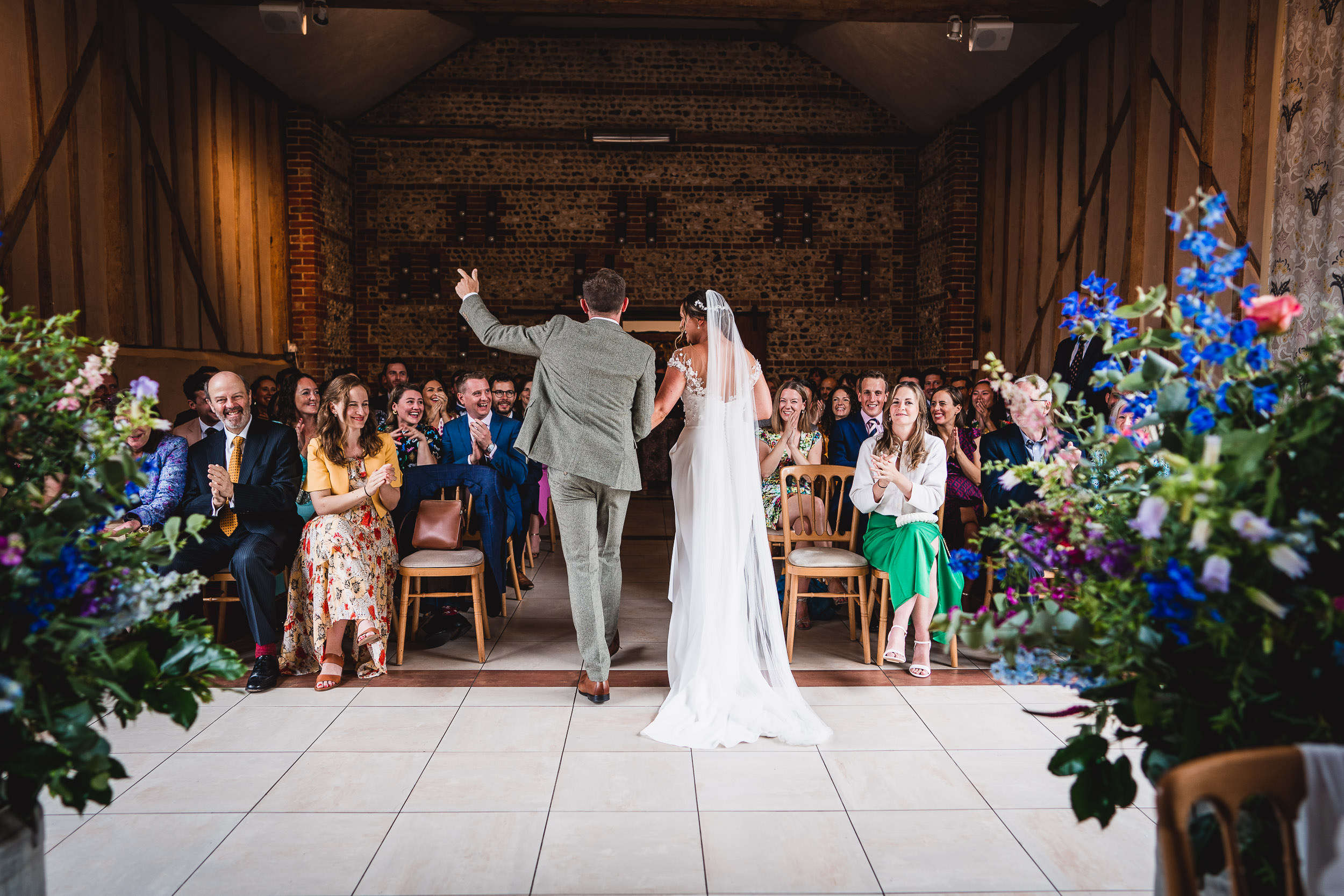 A bride and groom walk down the aisle, facing the audience, in a rustic venue decorated with colorful flowers. Guests are seated and clapping.