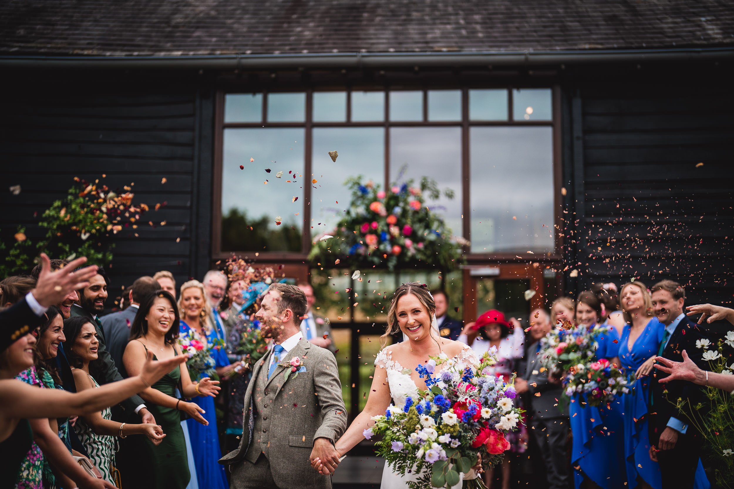 A bride and groom walk down an aisle outdoors, surrounded by guests throwing confetti. The couple smiles, holding hands and flowers. Guests are clapping and celebrating.