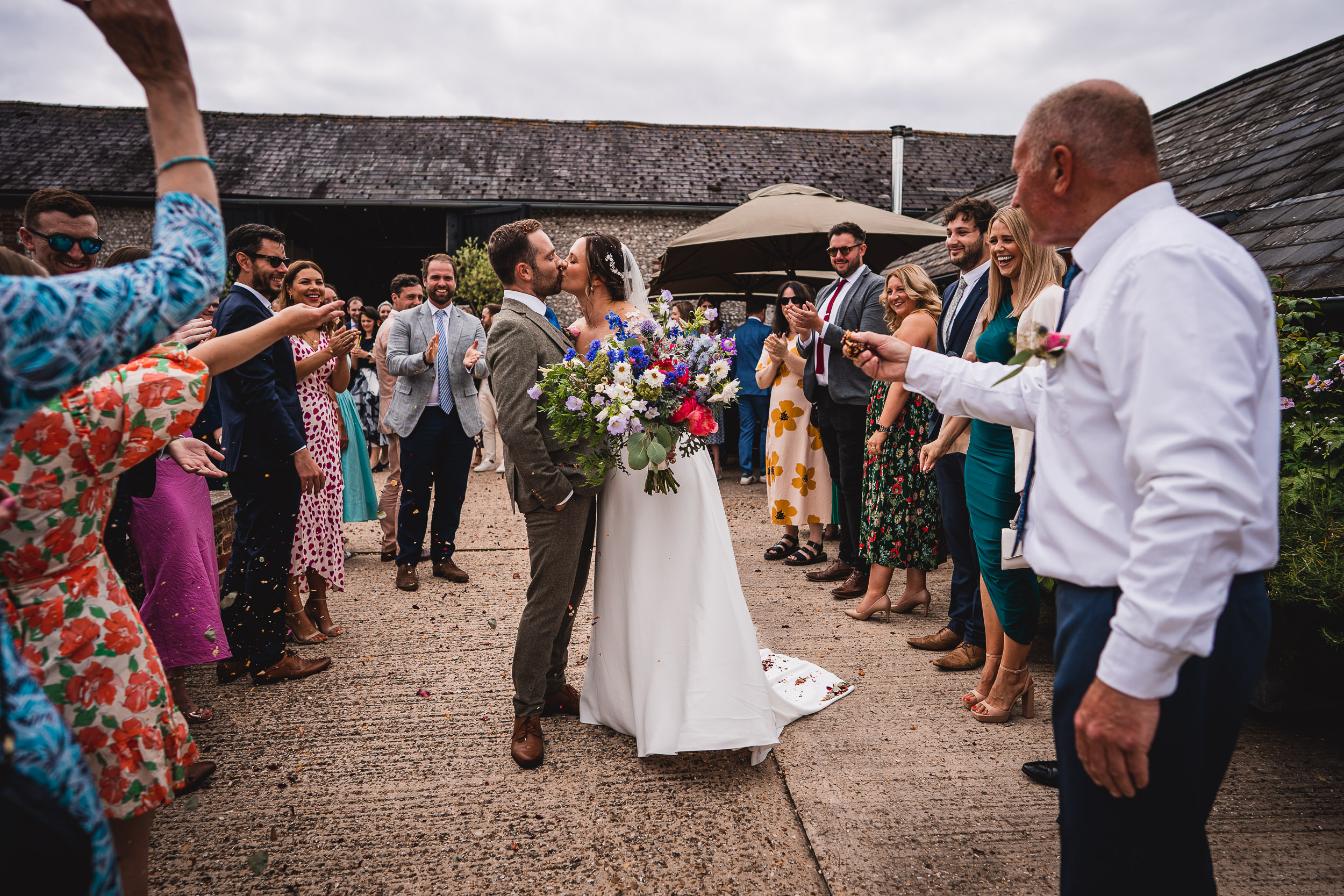 A couple kisses while surrounded by cheering guests at an outdoor wedding. The bride holds a bouquet of colorful flowers.