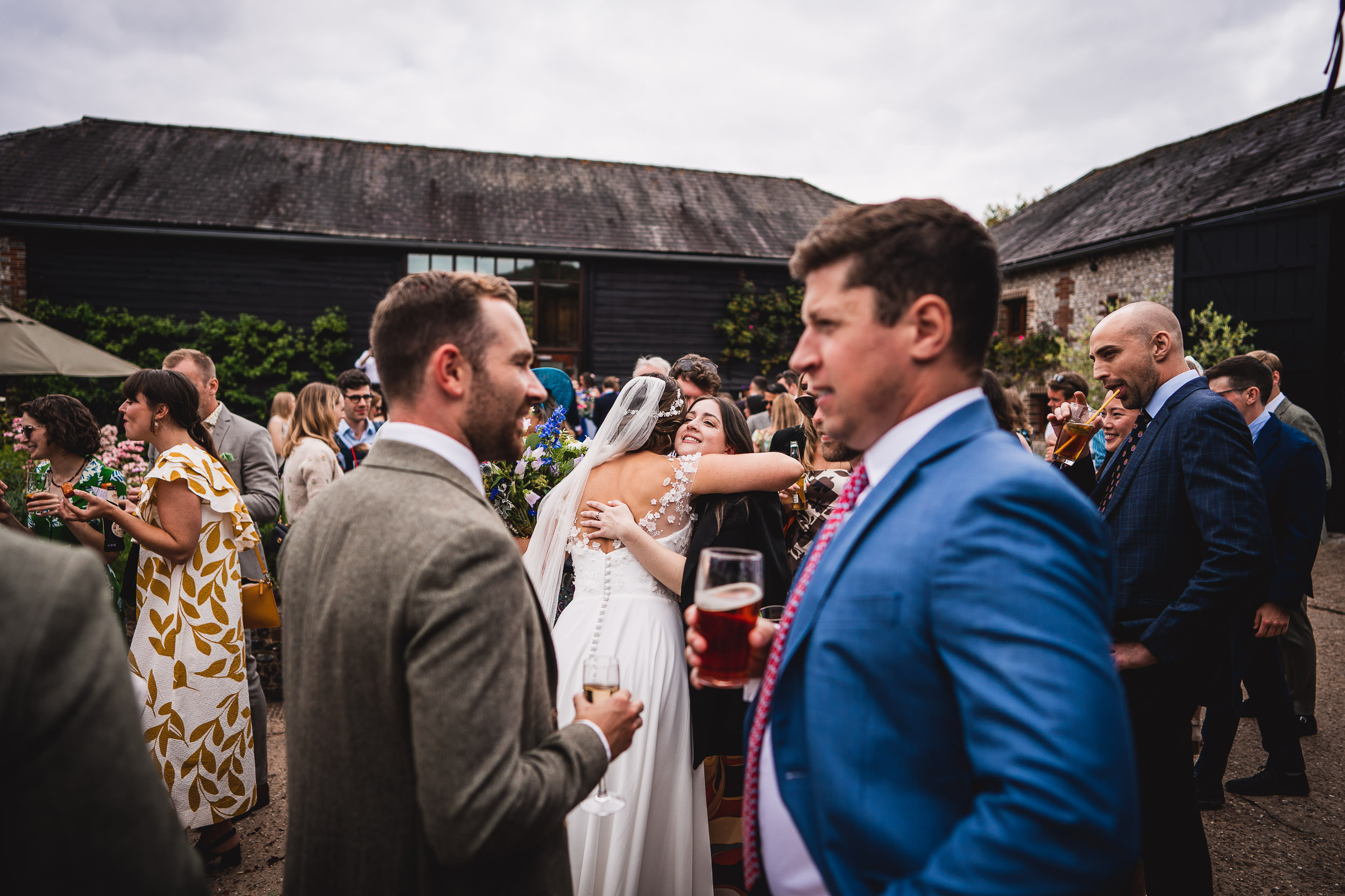 A wedding reception outdoors with guests mingling. A bride hugs a guest while others hold drinks and converse.