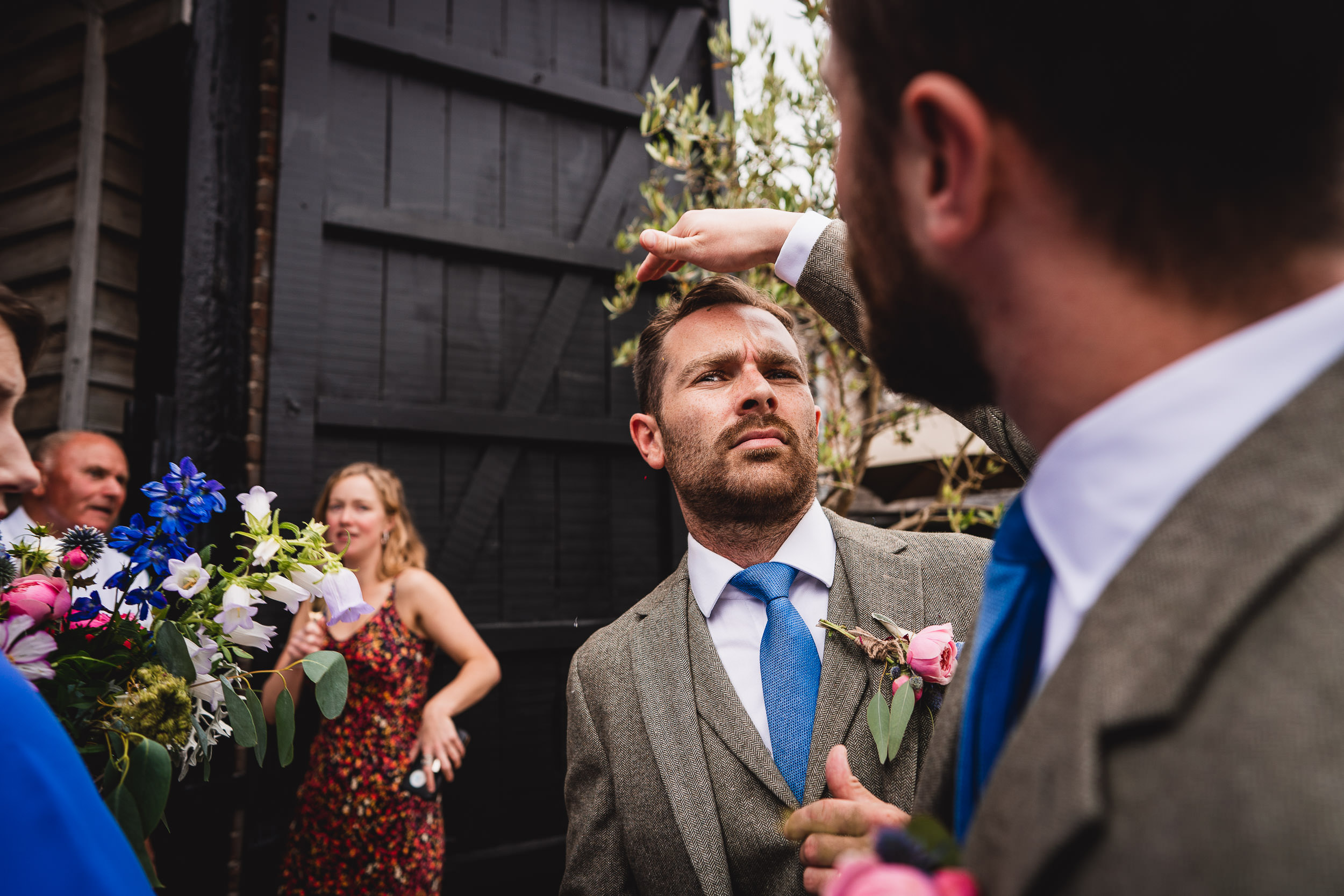 A groom in a grey suit with a blue tie is being adjusted by another man. People holding flowers are in the background.