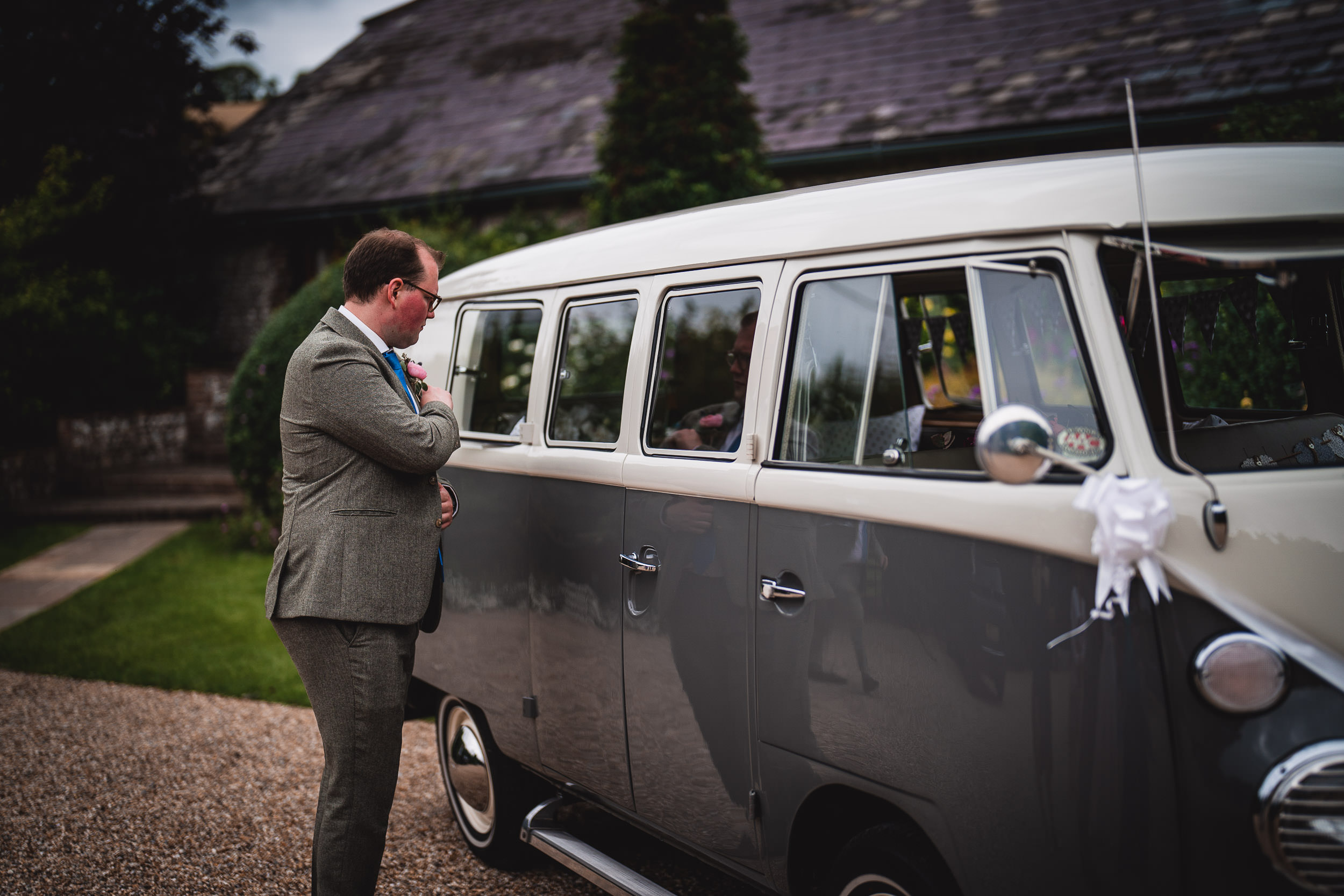 A man in a suit stands outside a vintage van, looking at its windows. The vehicle is gray and white, parked on a gravel path.