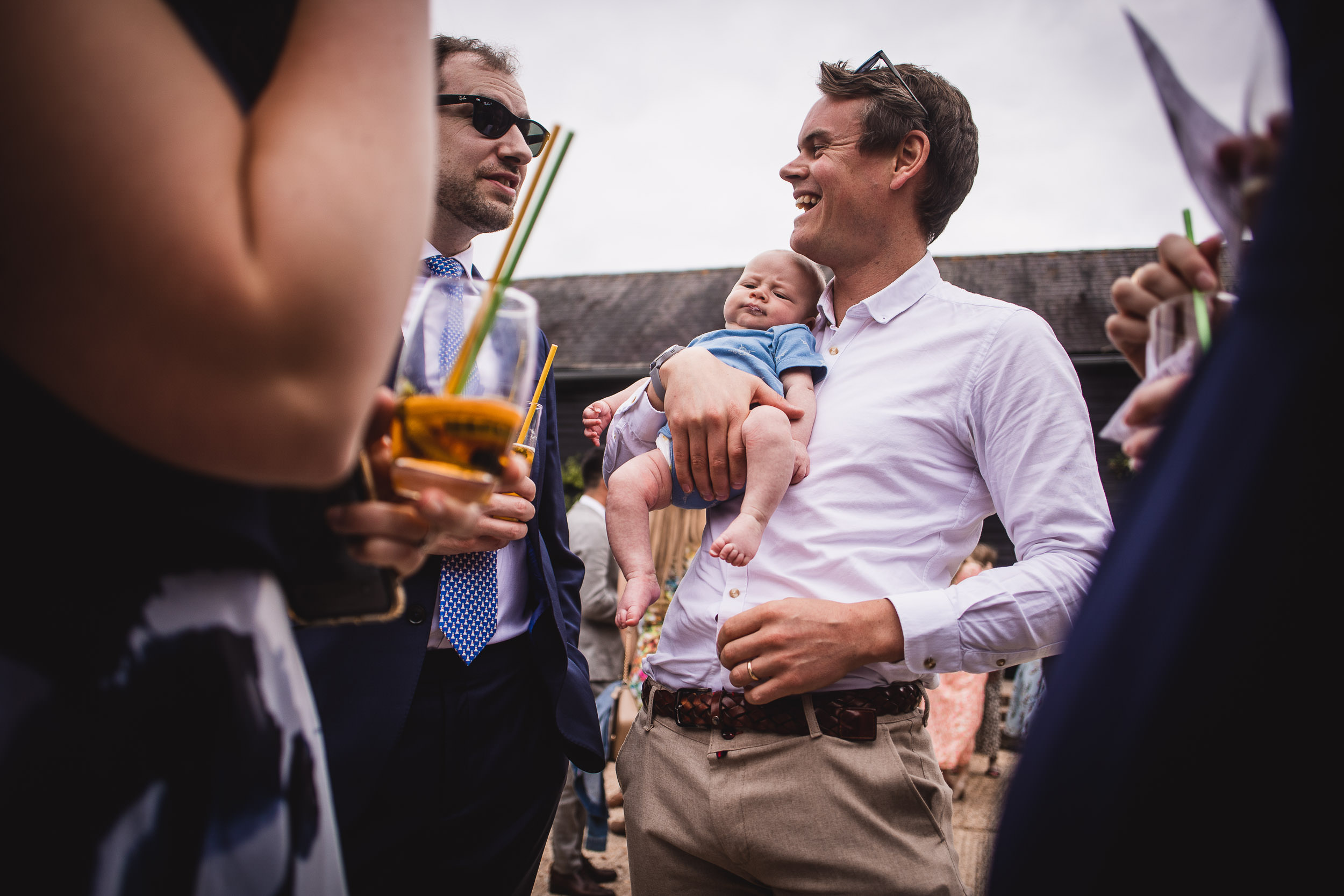 Two men, one holding a baby, chatting at an outdoor gathering. Others with drinks are partially visible around them.