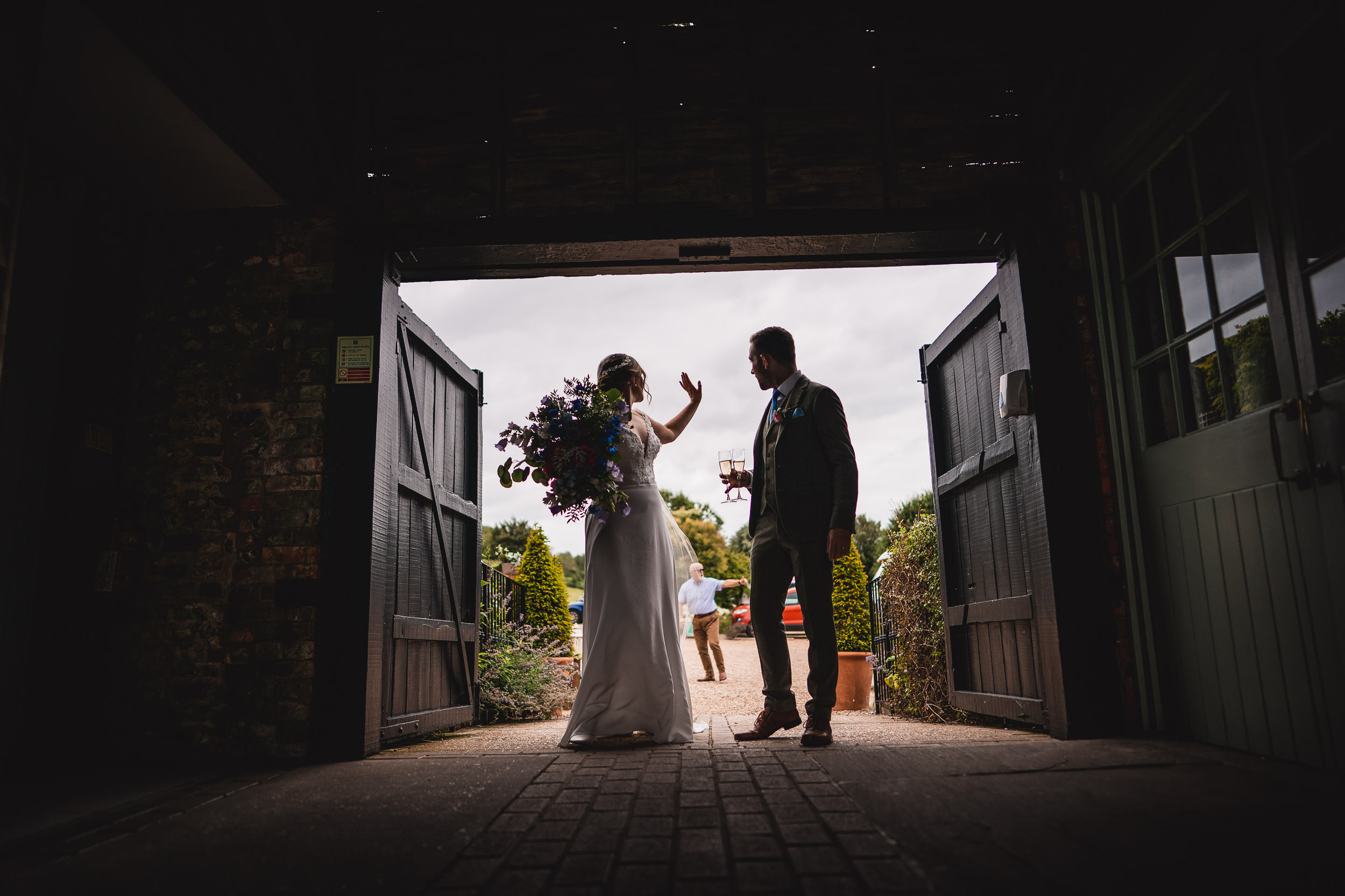 Bride and groom stand silhouetted in a doorway; the bride holds a large bouquet and raises her hand. A person walks in the background.