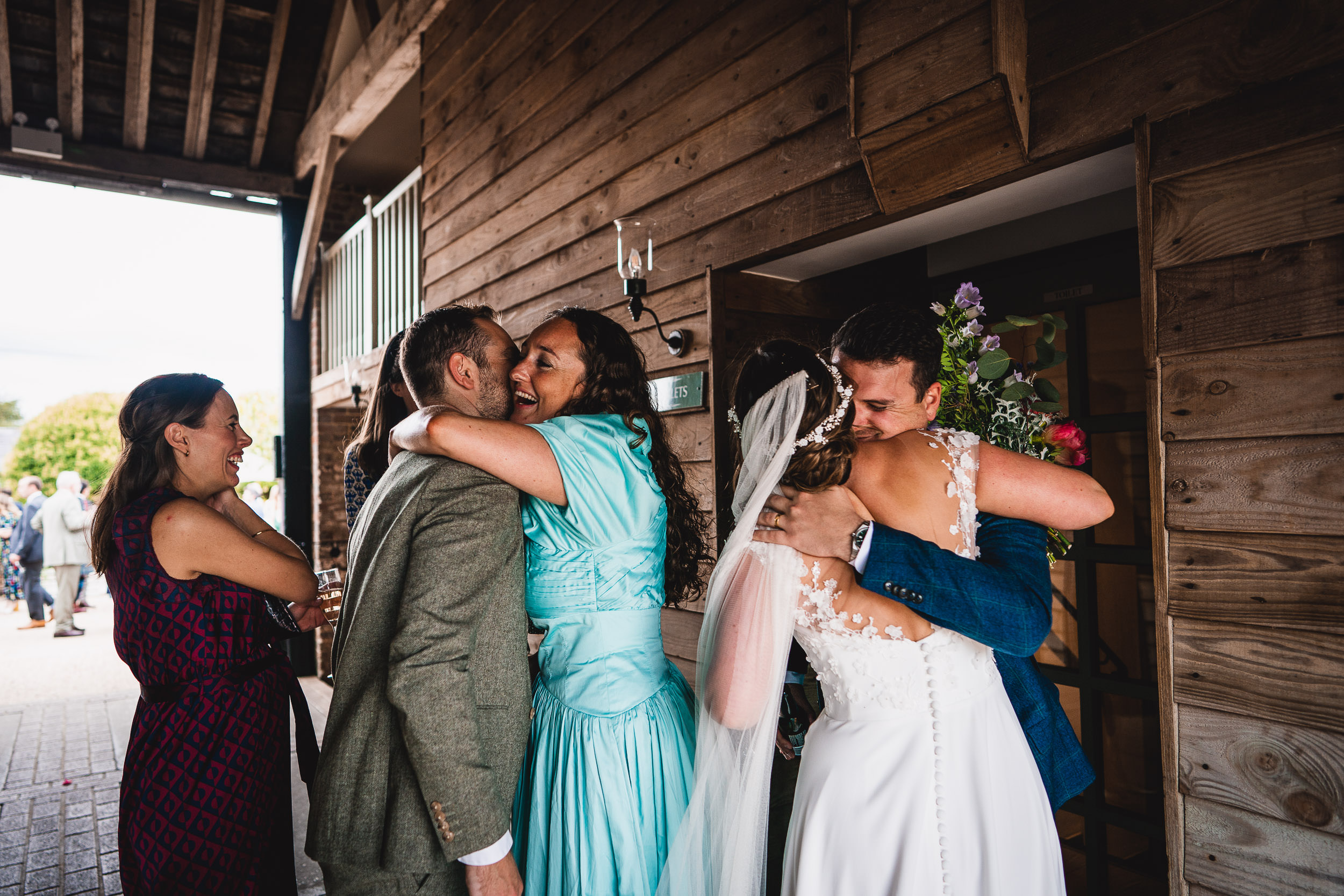 A bride and groom hug guests outside a wooden building during a celebration.