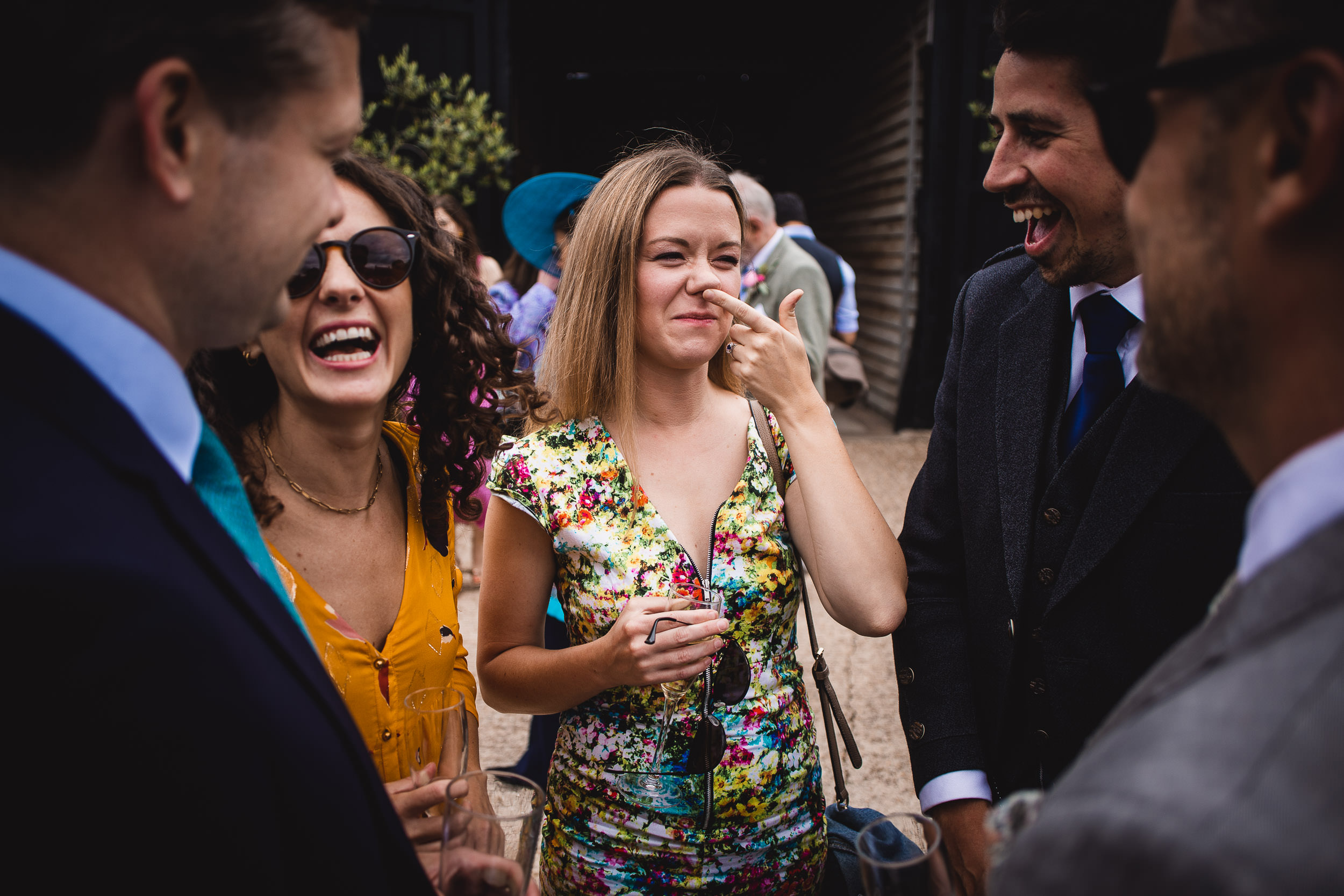 A group of people in formal attire are standing together outdoors, smiling and laughing. One woman is wearing a colorful floral dress and sunglasses are held in her hand.