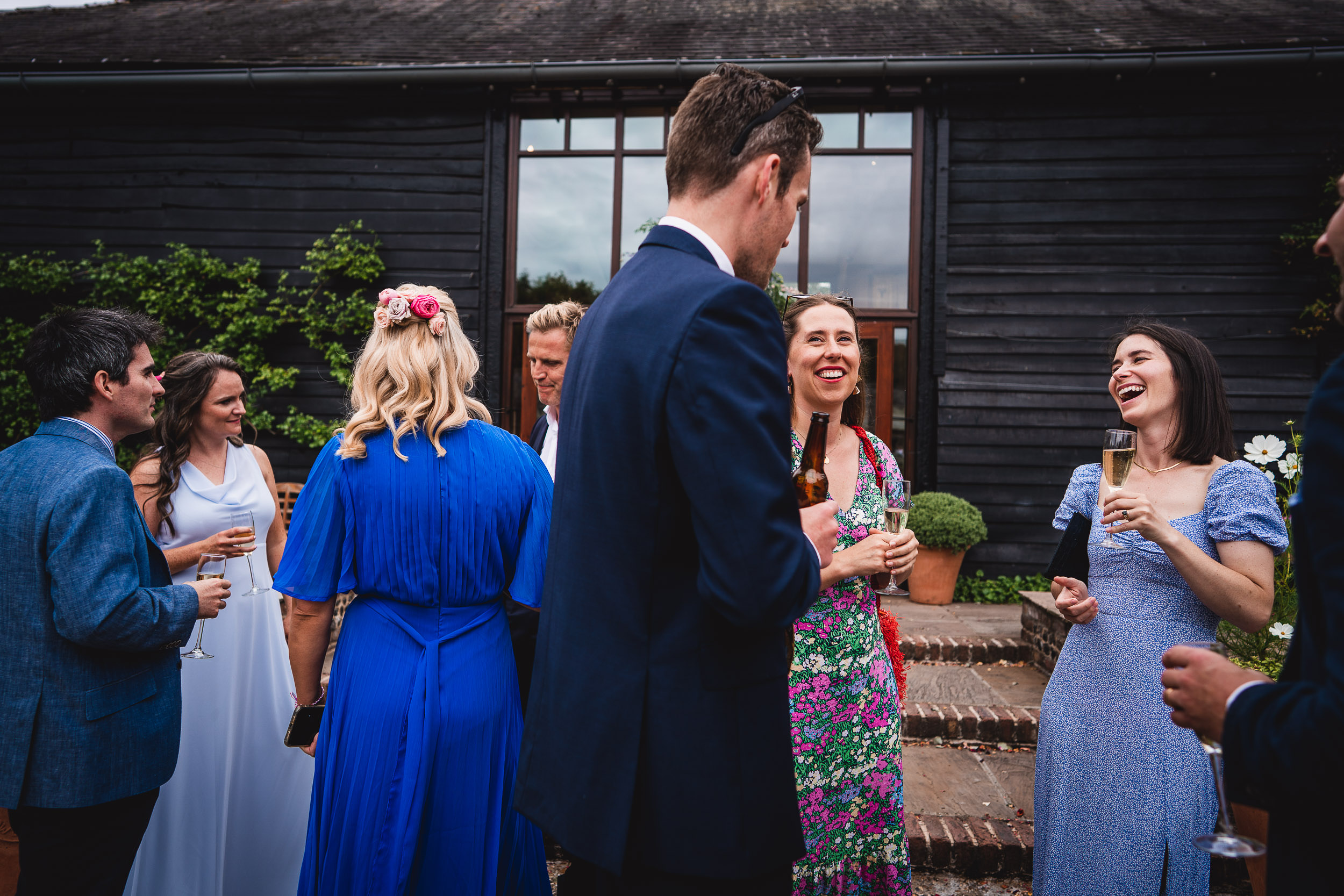 A group of people in formal attire stand outdoors, smiling and talking, holding drinks. A dark wooden building is in the background.