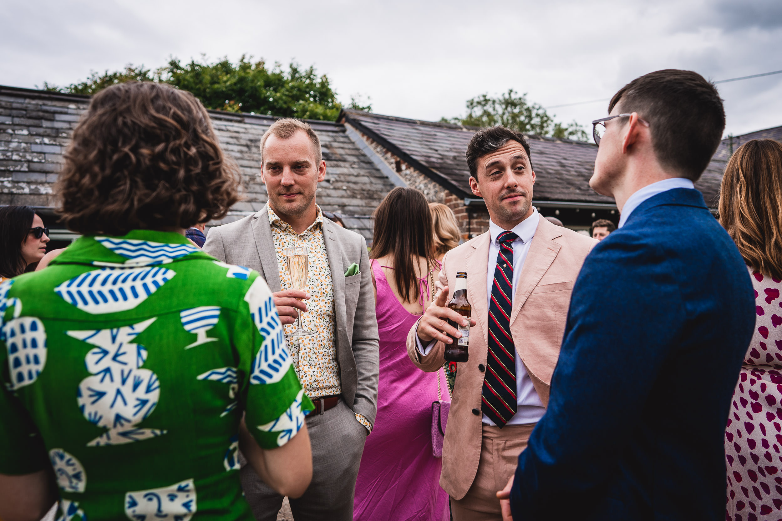 A group of four people in formal attire converse outdoors at a social event. One person holds a drink.