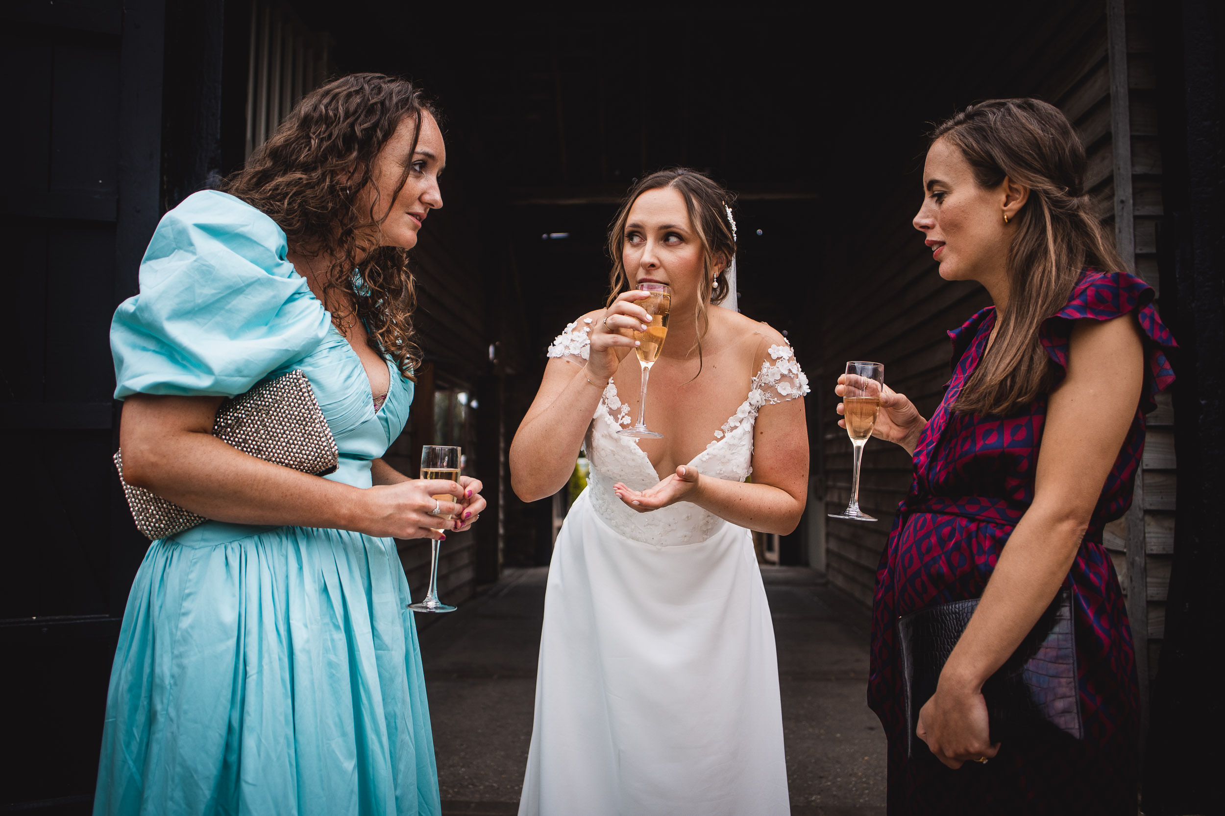 Three women stand together, holding glasses of champagne. The woman in the center, wearing a white dress, is sipping her drink. They are in a dimly lit outdoor setting.