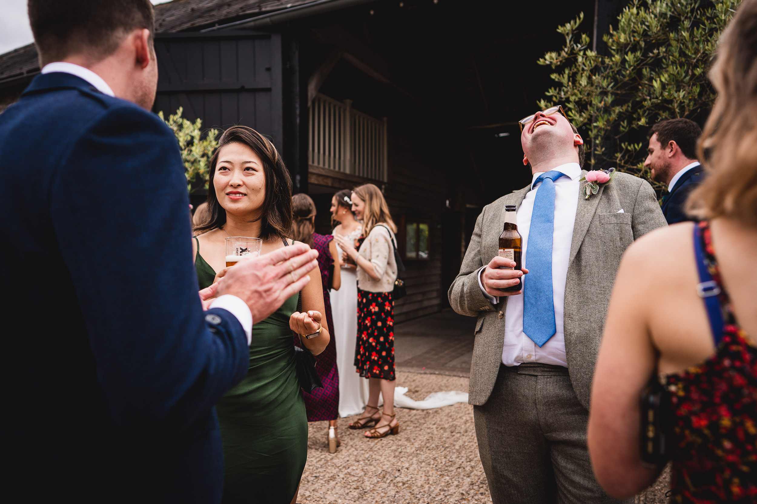 A group of people at an outdoor gathering, dressed in formal attire, are conversing and laughing. One man is holding a beer bottle.