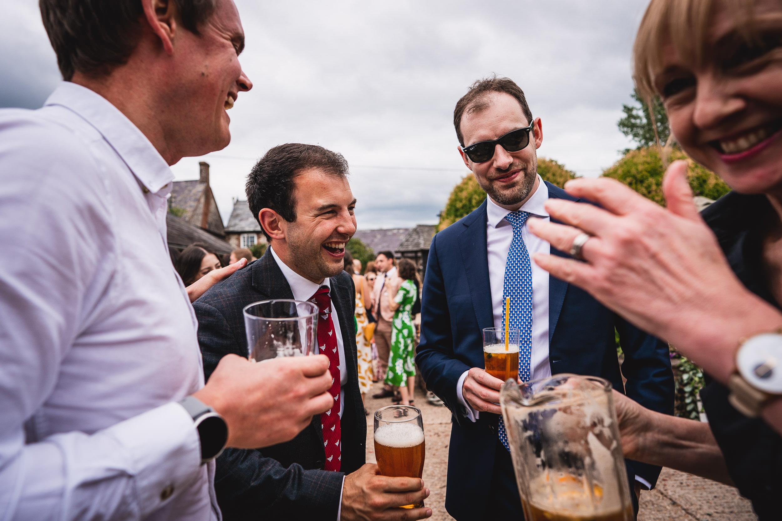 Group of people in formal attire enjoying drinks outdoors, with two men holding glasses of beer and a woman pouring from a pitcher.