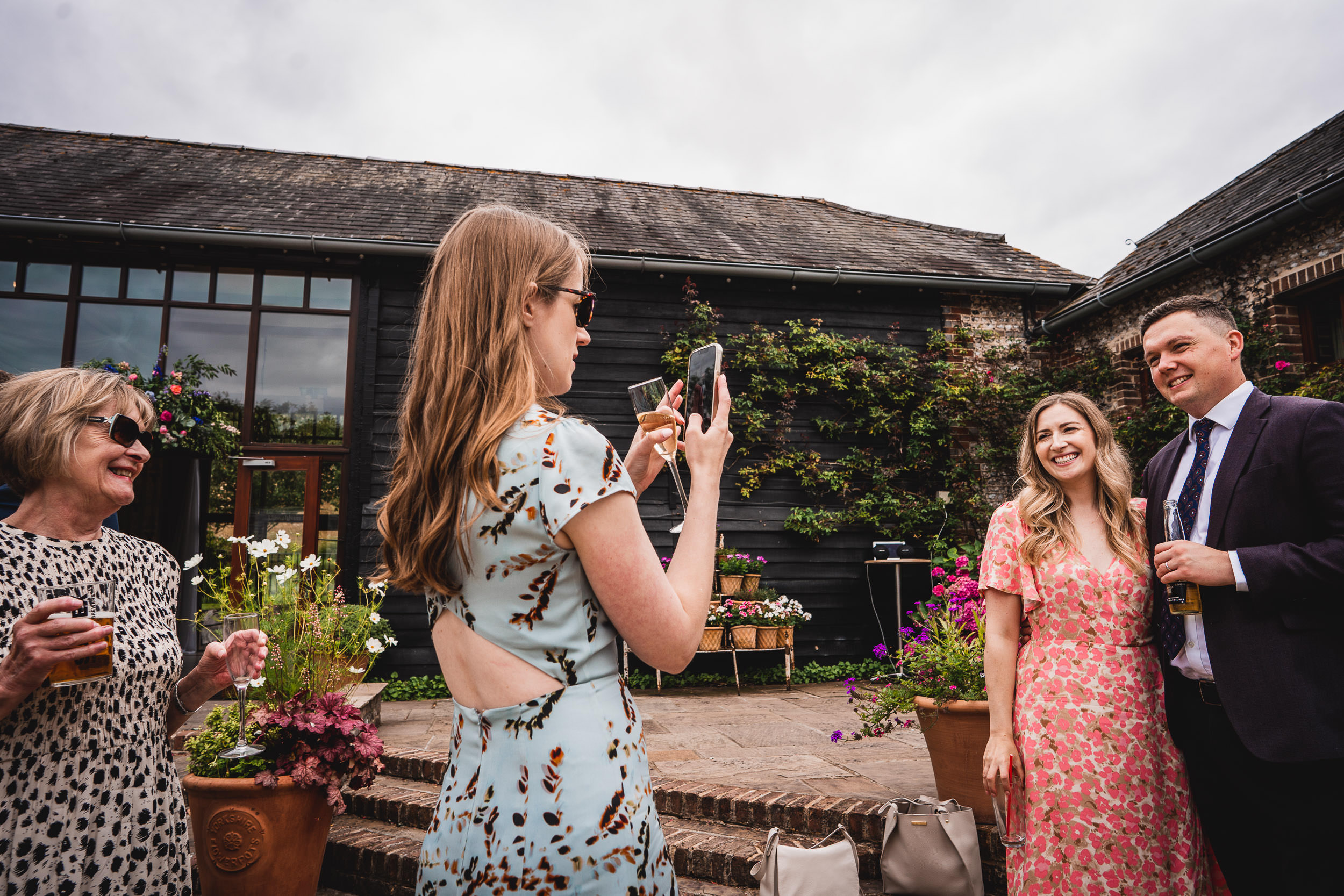 A woman takes a photo of a smiling couple at an outdoor gathering, surrounded by flowers and a building with large windows.