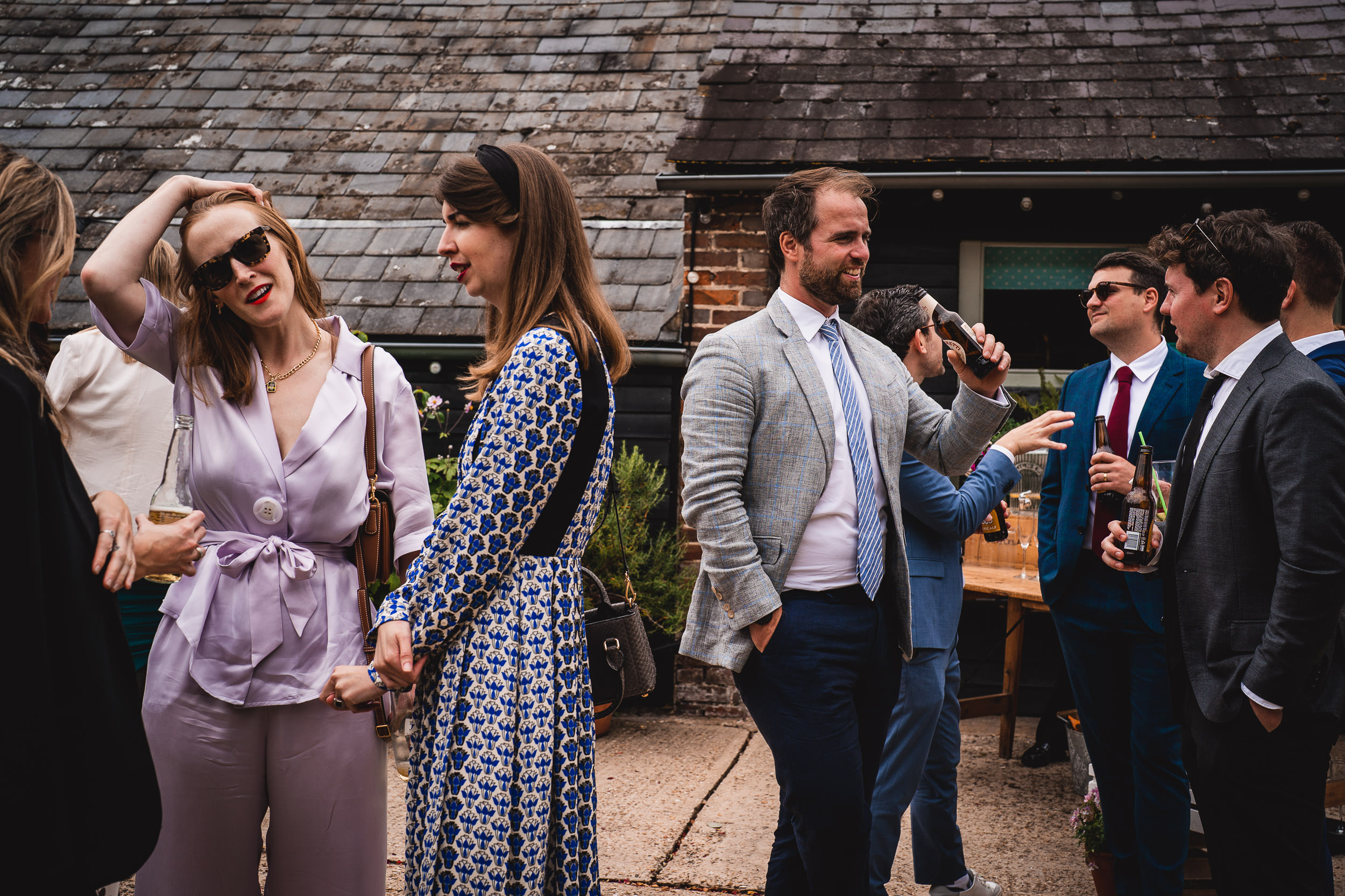 A group of people in formal attire conversing outdoors. A man holds a microphone, while others hold drinks. There is a building with a tiled roof in the background.